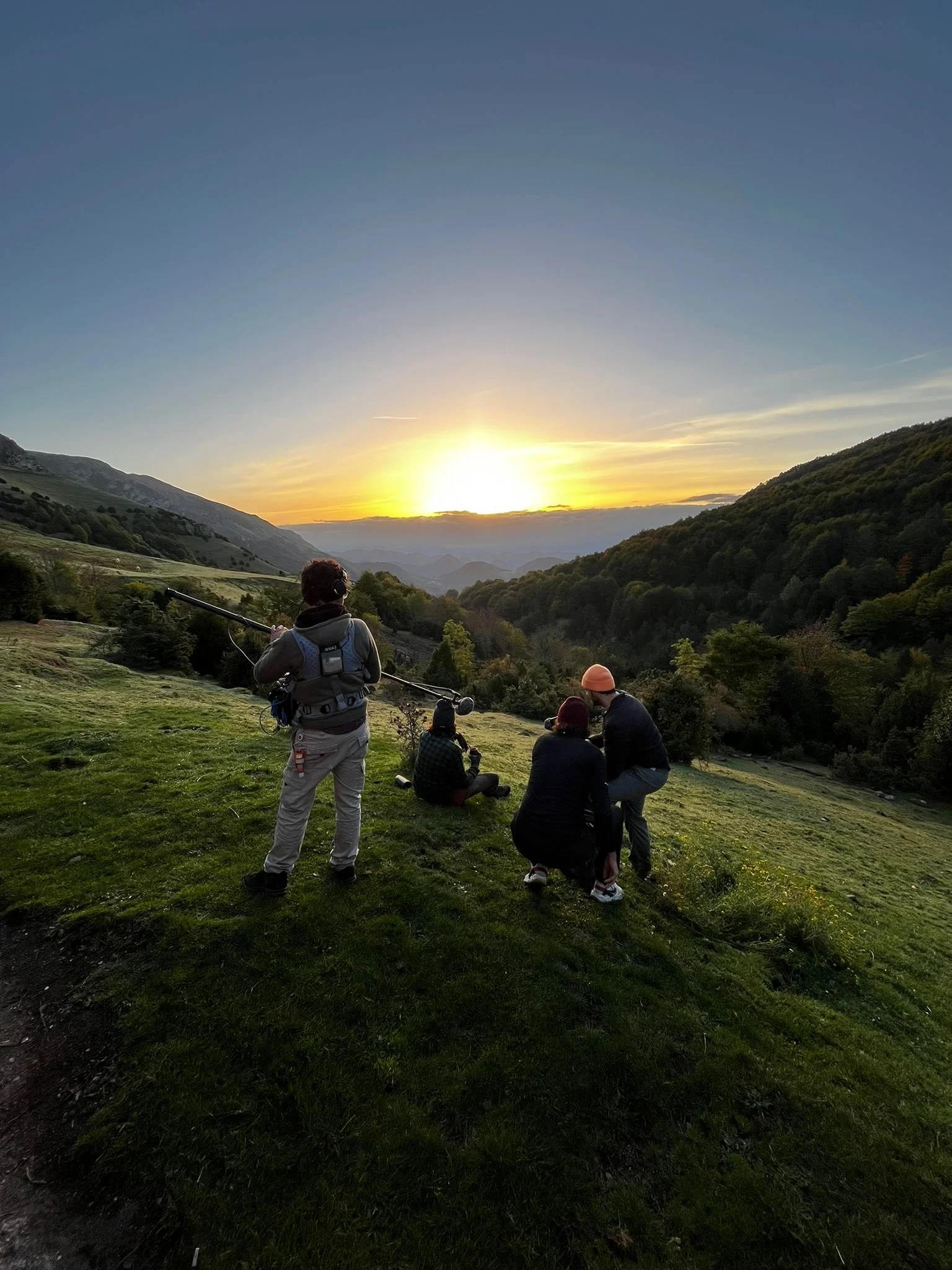 A group of four people enjoying a sunset in a mountainous landscape, with lush green trees and hills.