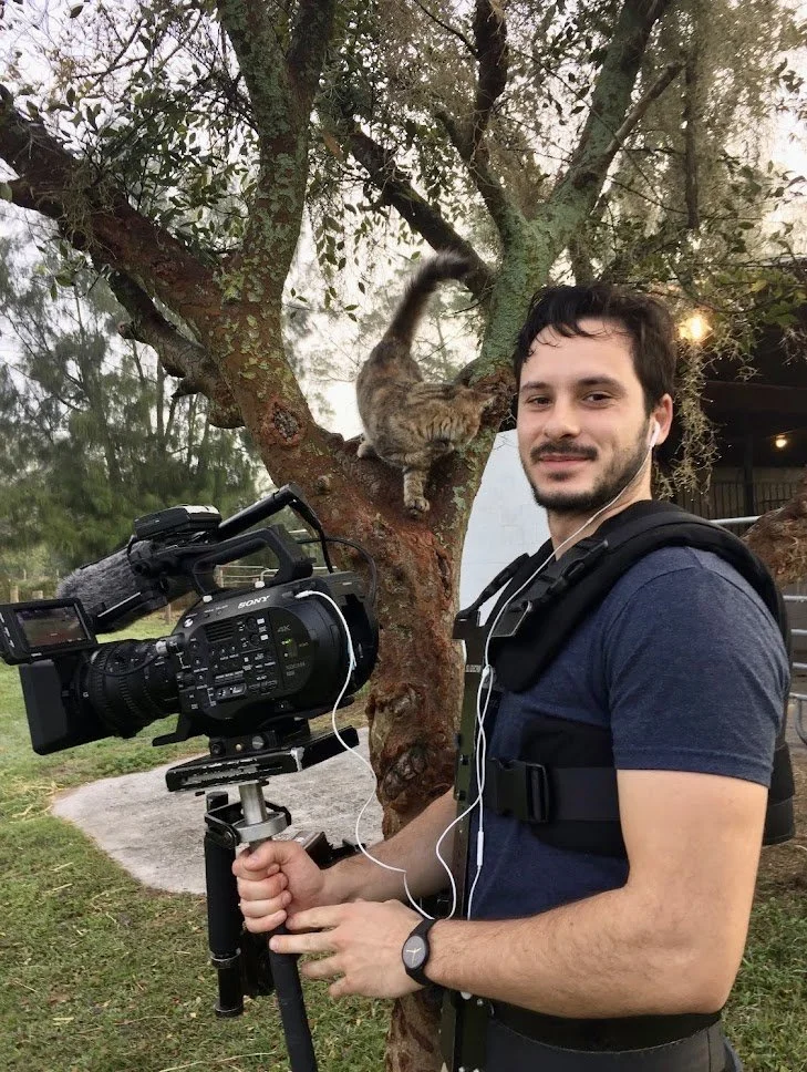 A man with dark hair, a beard, and a grey watch holding a professional video camera on a tripod in an outdoor setting. In the background, a cat is climbing a tree with green leaves.