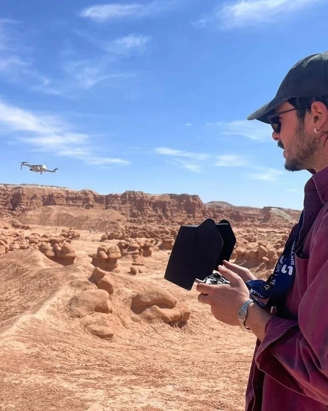A man wearing sunglasses, a dark cap, and a maroon jacket operates a drone controller in a desert landscape with rock formations. A drone is flying in the blue sky in the background.