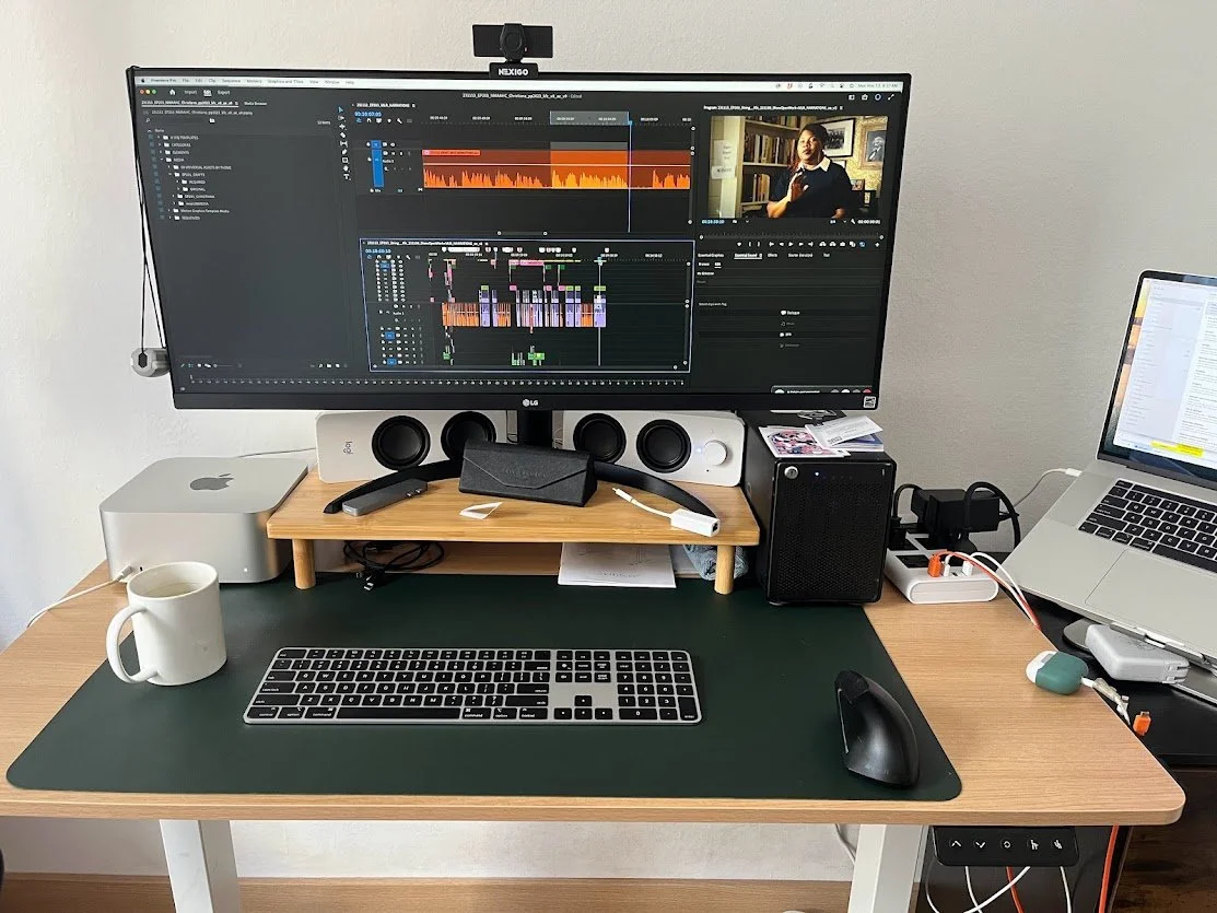 A computer desk with a large monitor displaying video editing software, a laptop, a keyboard, a mouse, and various speakers and accessories on the desk and shelves.