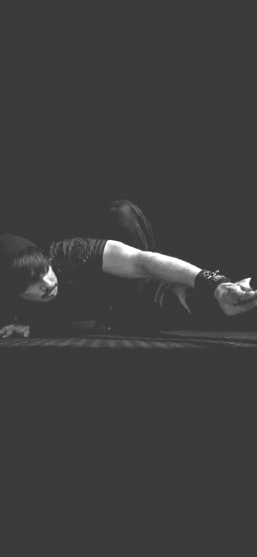 A person lying on their side on a dark surface, reaching out with their arm, wearing a dark, long-sleeved shirt and black wrist accessories, with dark hair and a focused expression.