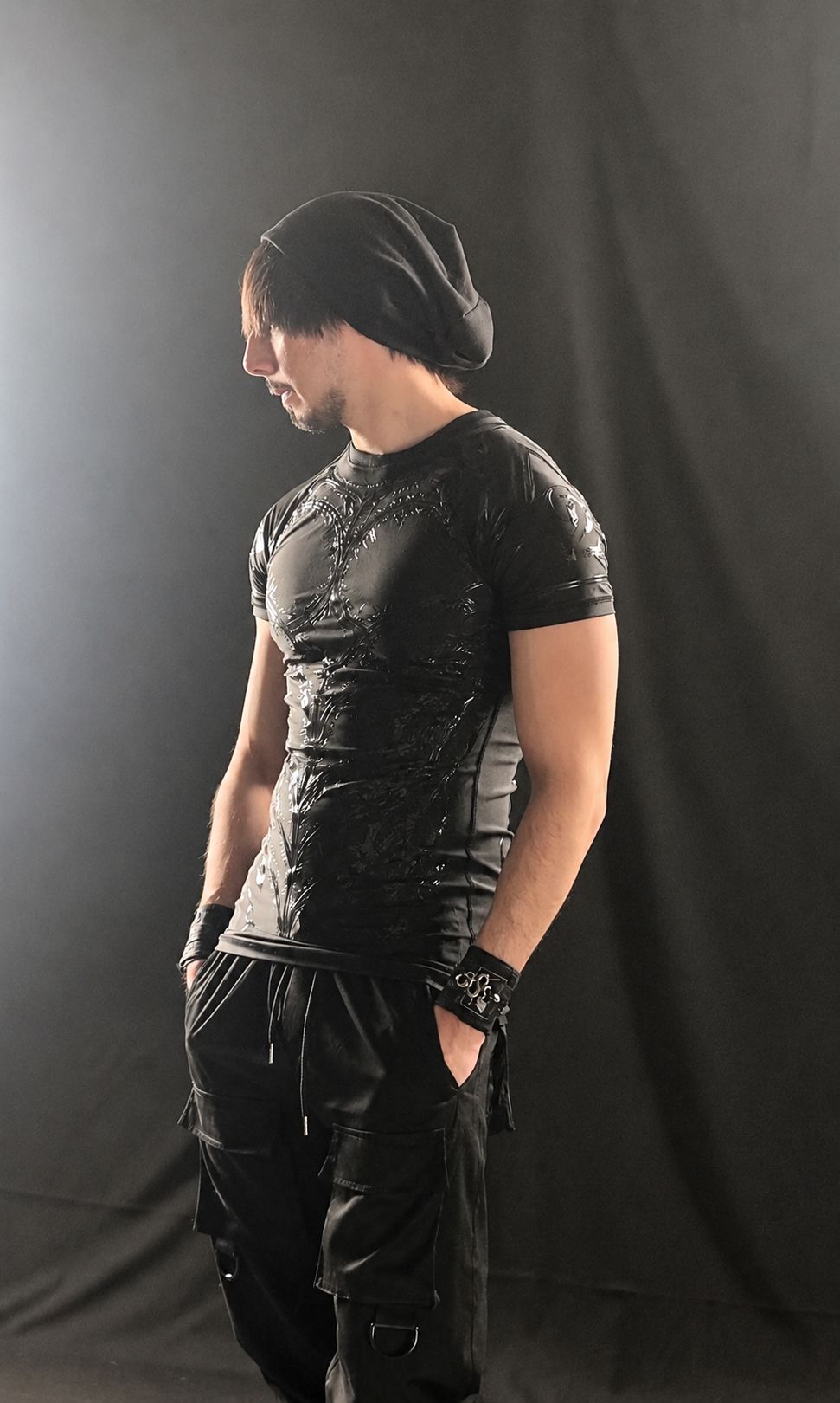 A man stands sideways wearing a black shiny short-sleeve shirt, baggy cargo pants, a black wristband, and a black beanie against a dark background.
