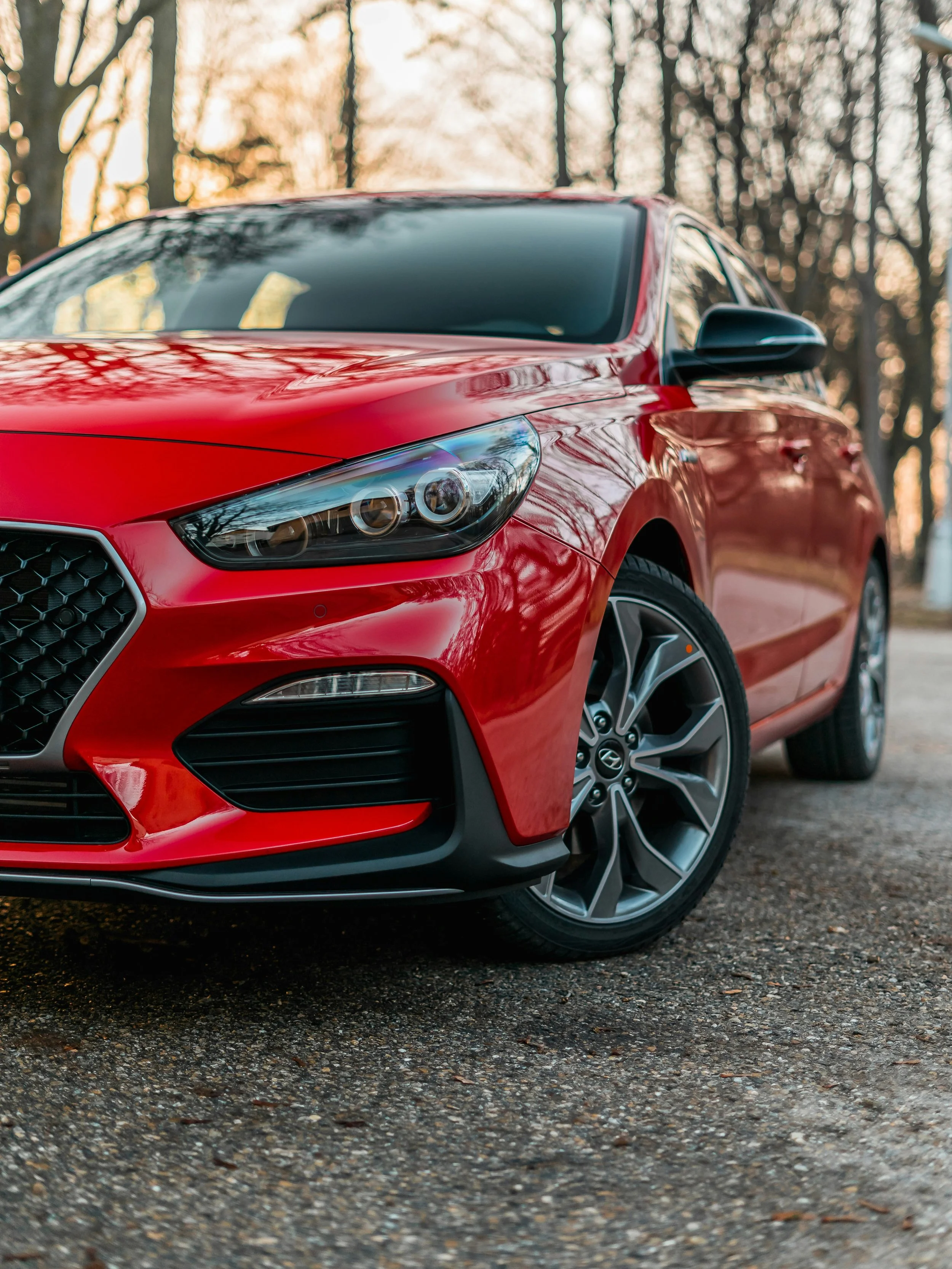 Close-up of the front and side of a red car parked outdoors, with trees and sunset in the background.
