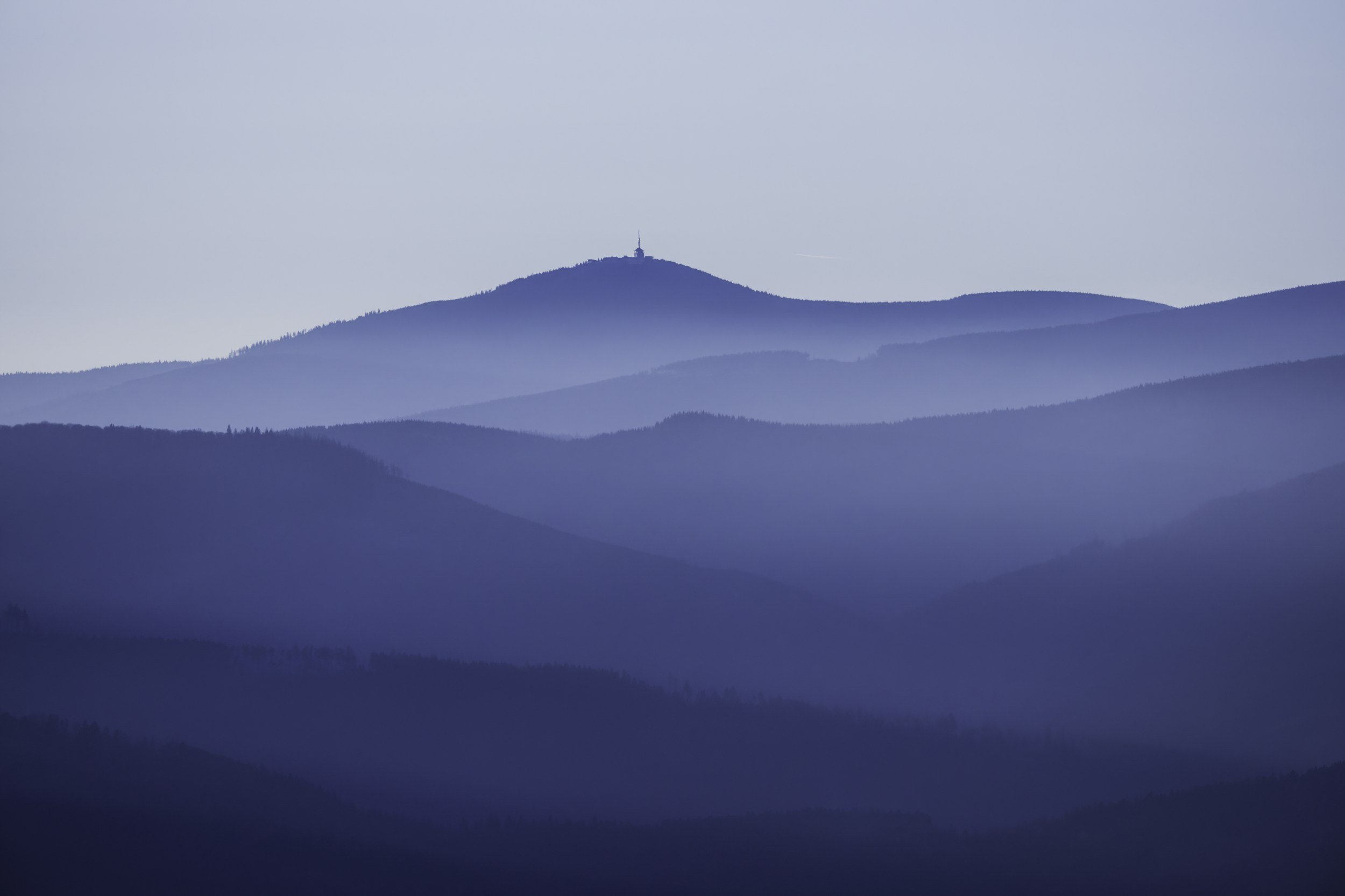 A mountain with a tower or antenna at the summit, surrounded by layered blue and green mountain ranges and a clear sky.