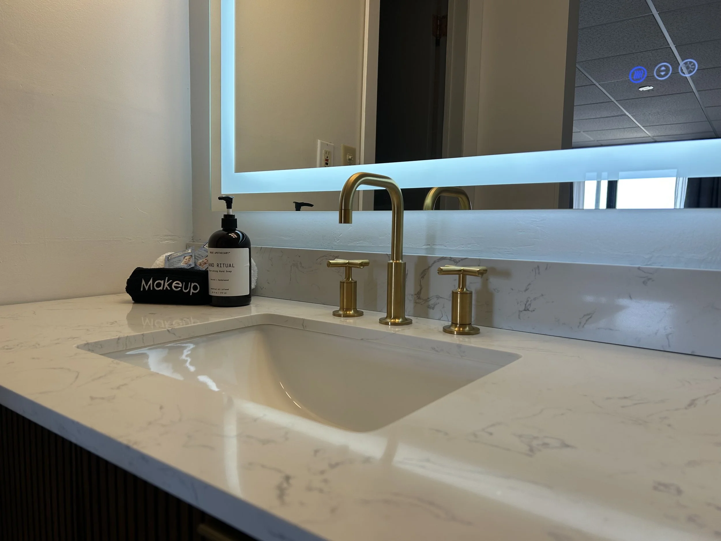 Bathroom sink with a white marble countertop, gold faucet, a bottle of hand soap, a black makeup bag, and a large mirror with blue LED backlighting. There are electrical outlets and a dark doorway in the background.