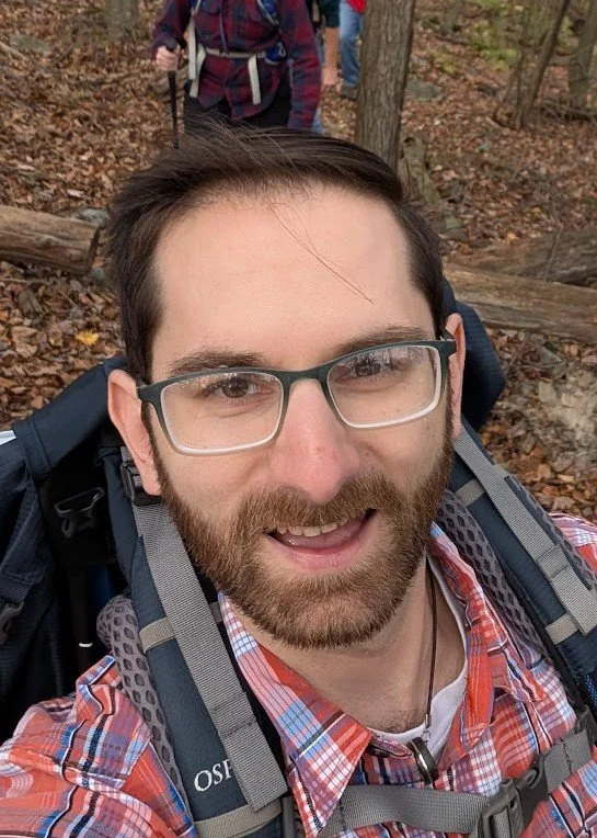 Close-up of a smiling man with glasses, beard, wearing a plaid shirt and carrying a backpack, in a wooded outdoor setting during daytime.