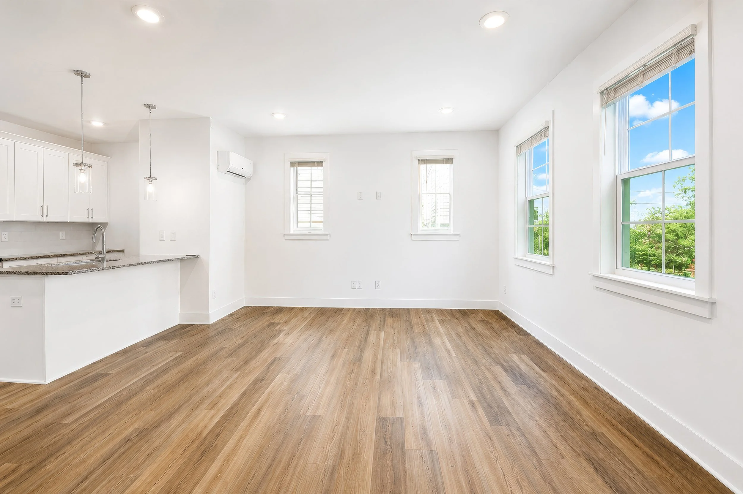 Empty room with wooden flooring, white walls, multiple windows with blinds, and a ceiling with recessed lighting. Part of an open kitchen area with white cabinets and pendant lights is visible on the left.