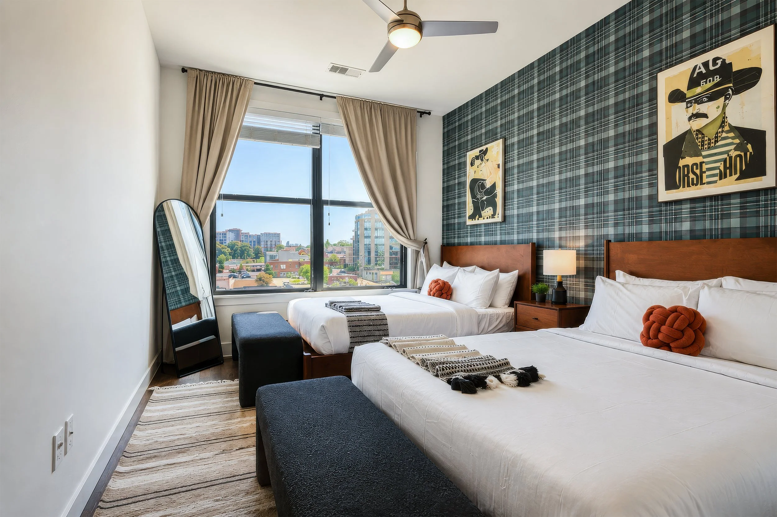 A modern hotel room featuring two beds with white bedding, decorative pillows, a nightstand with a lamp and plant, large window with beige curtains, and wall art with a plaid pattern behind the beds.