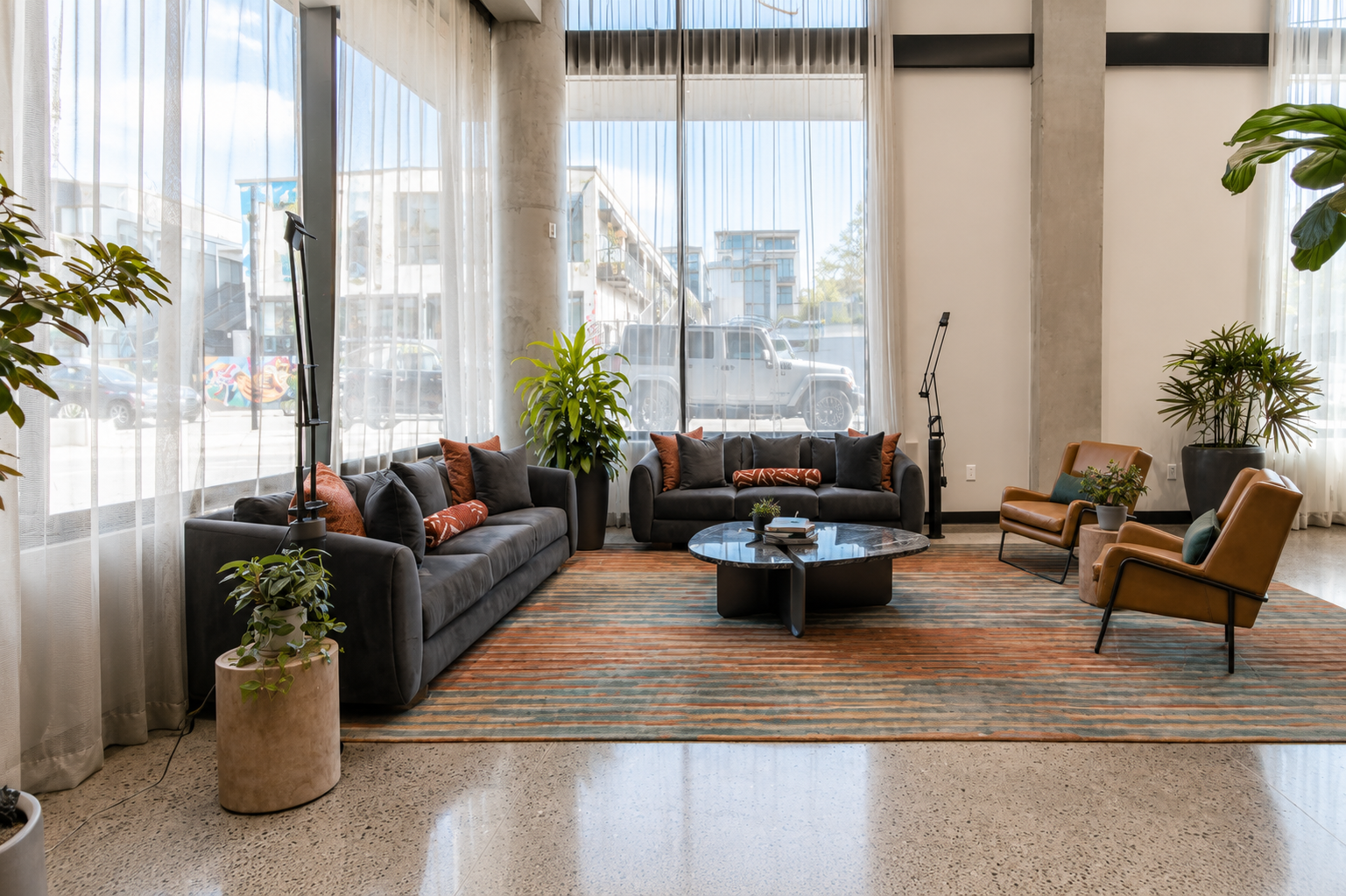 Modern hotel lobby with black leather sofas, orange and black throw pillows, a circular marble coffee table, and armchairs, surrounded by large indoor plants and floor-to-ceiling windows with sheer curtains.