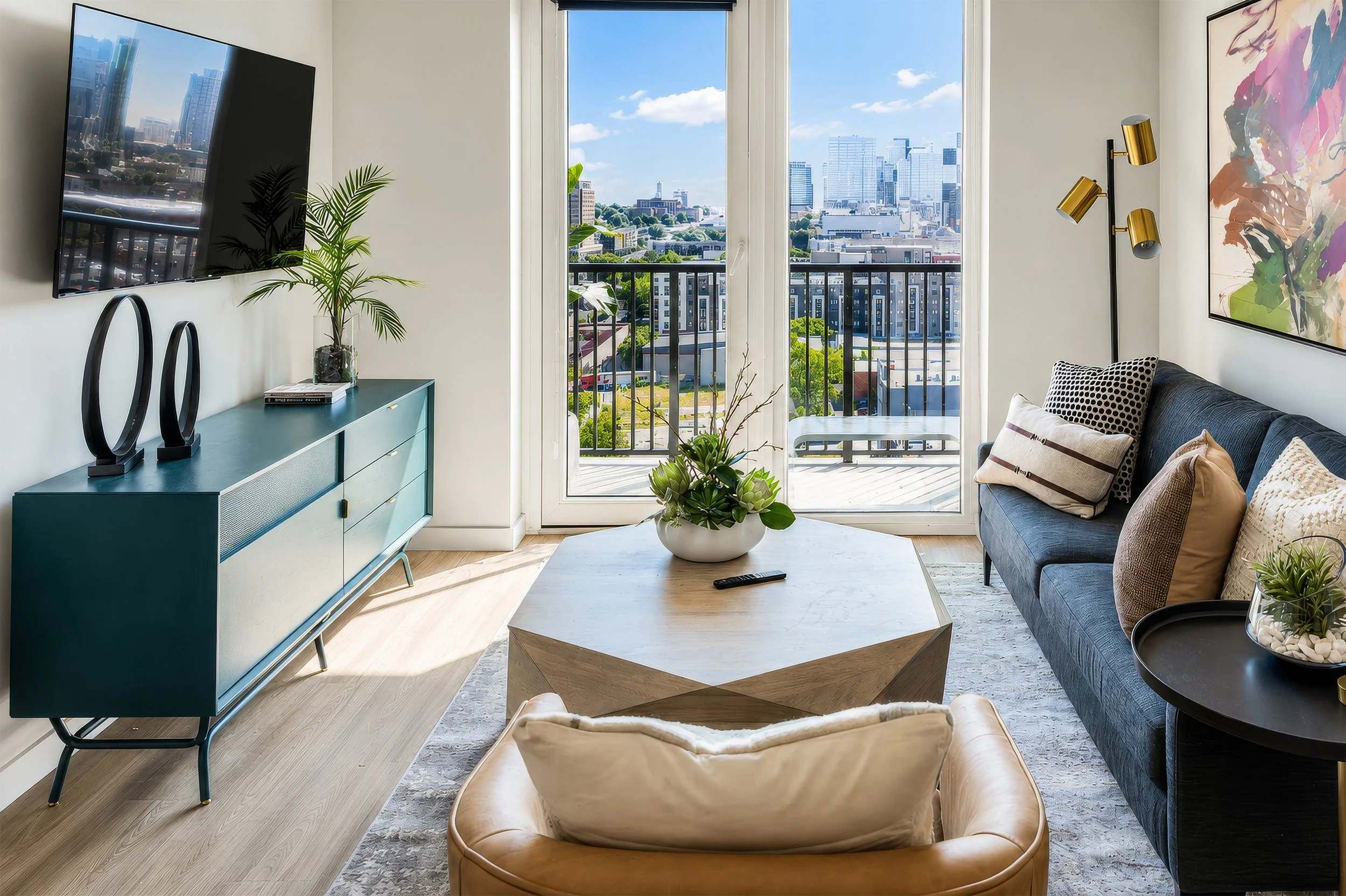 Modern living room with large window overlooking city skyline, with a black sofa, decorative pillows, a coffee table, a teal sideboard with sculptures, indoor plants, a mounted flat-screen TV, and abstract wall art.