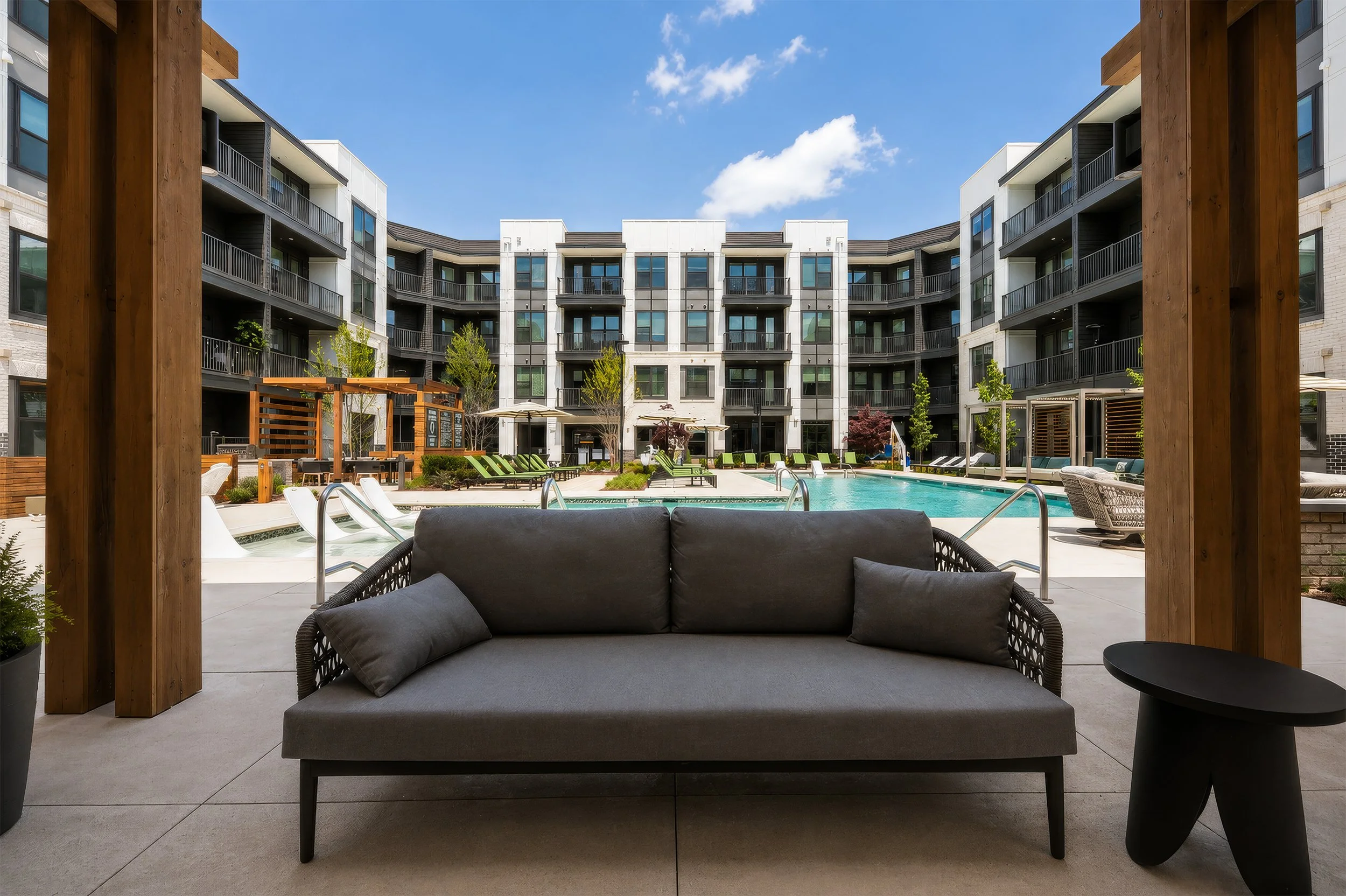 A view from a shaded patio seating area looking out at a swimming pool with lounge chairs, trees, and apartment buildings in the background against a blue sky with clouds.