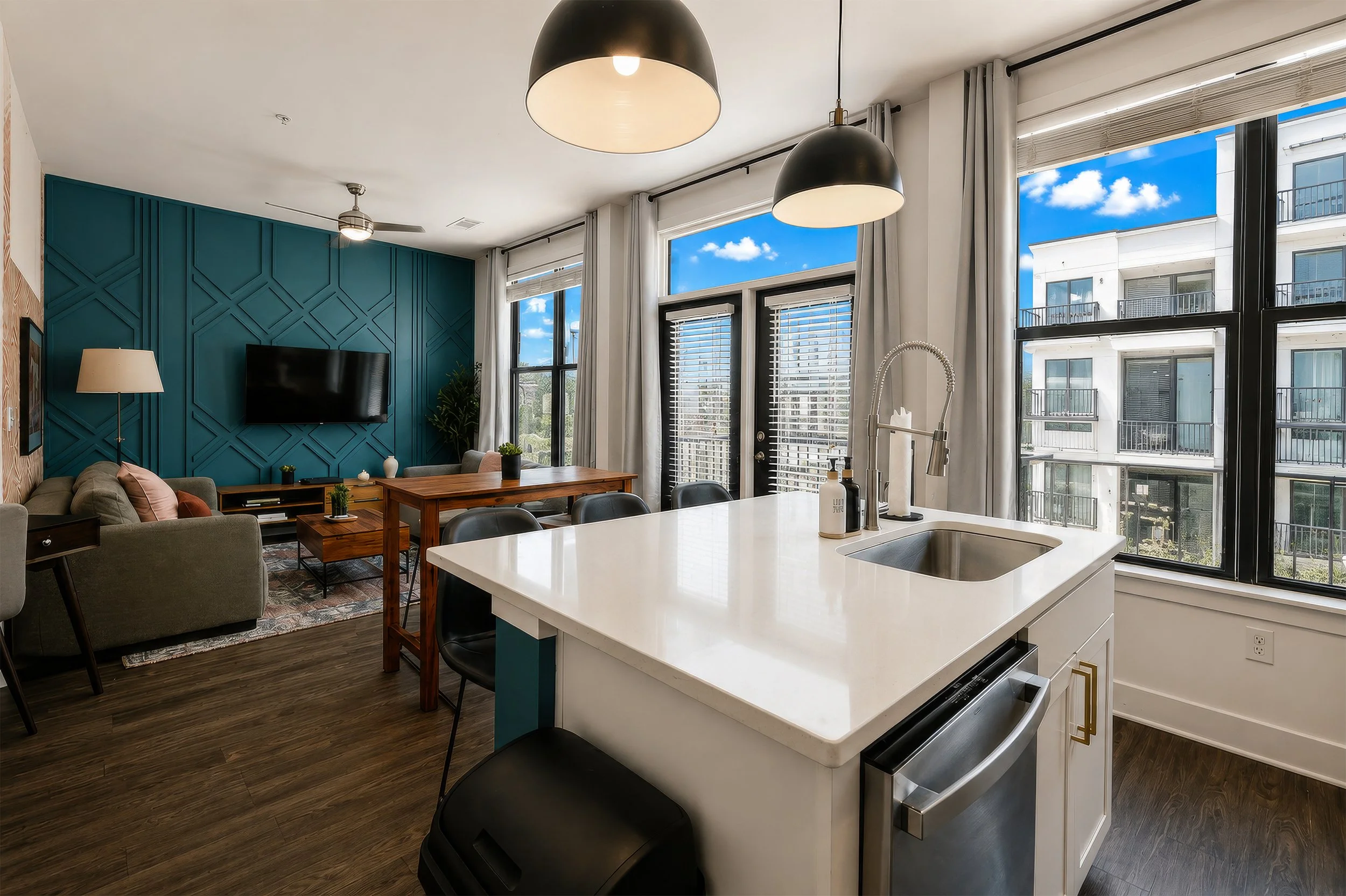Modern living room with a blue accent wall, TV, sofa, and large windows showing a cityscape with clouds and adjacent apartment balconies. Kitchen counters with a sink and black stools.
