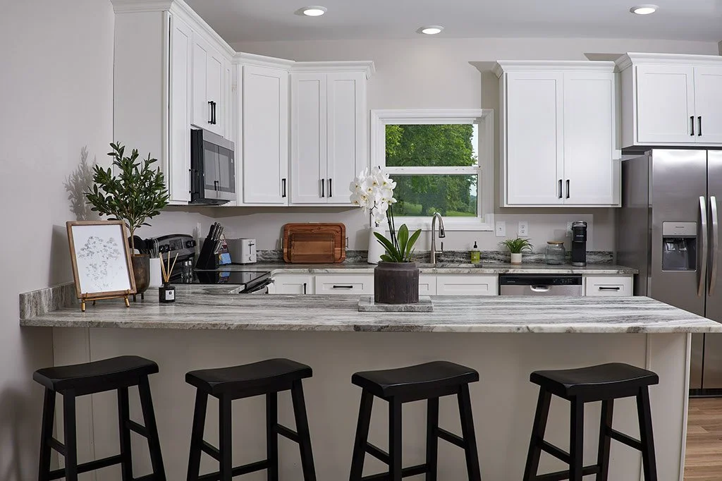 Modern white kitchen with granite countertops, stainless steel appliances, and black bar stools. A central counter features a potted orchid, with a window showing green trees outside.