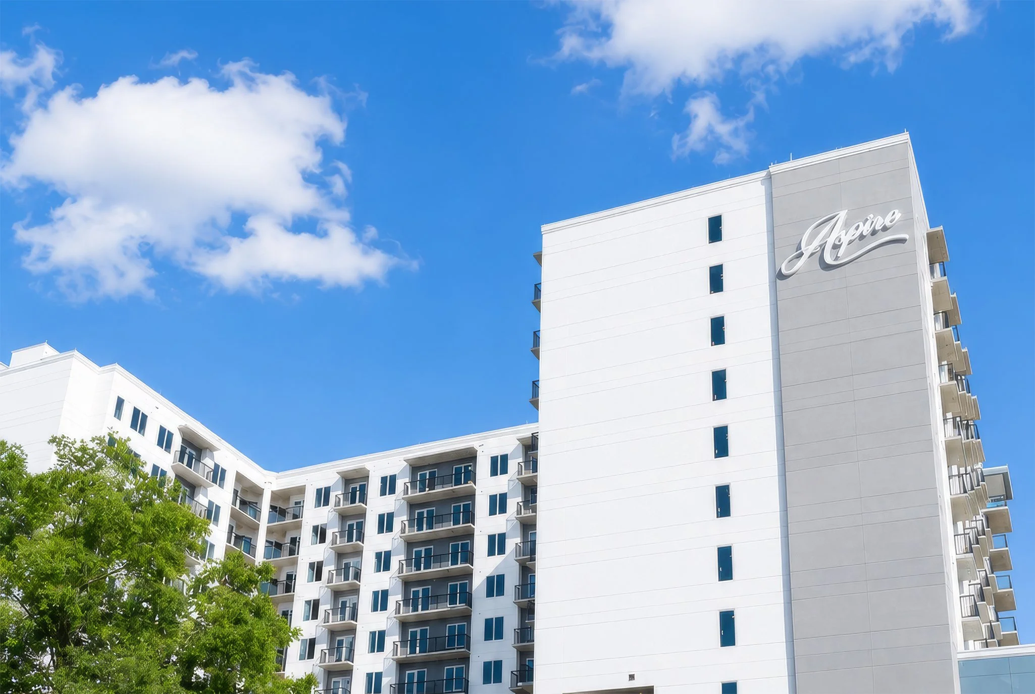 A tall white modern apartment building with multiple balconies and a large sign that says 'Aprea' on the upper part of the building. The sky is bright blue with scattered clouds, and there are green trees in the foreground.