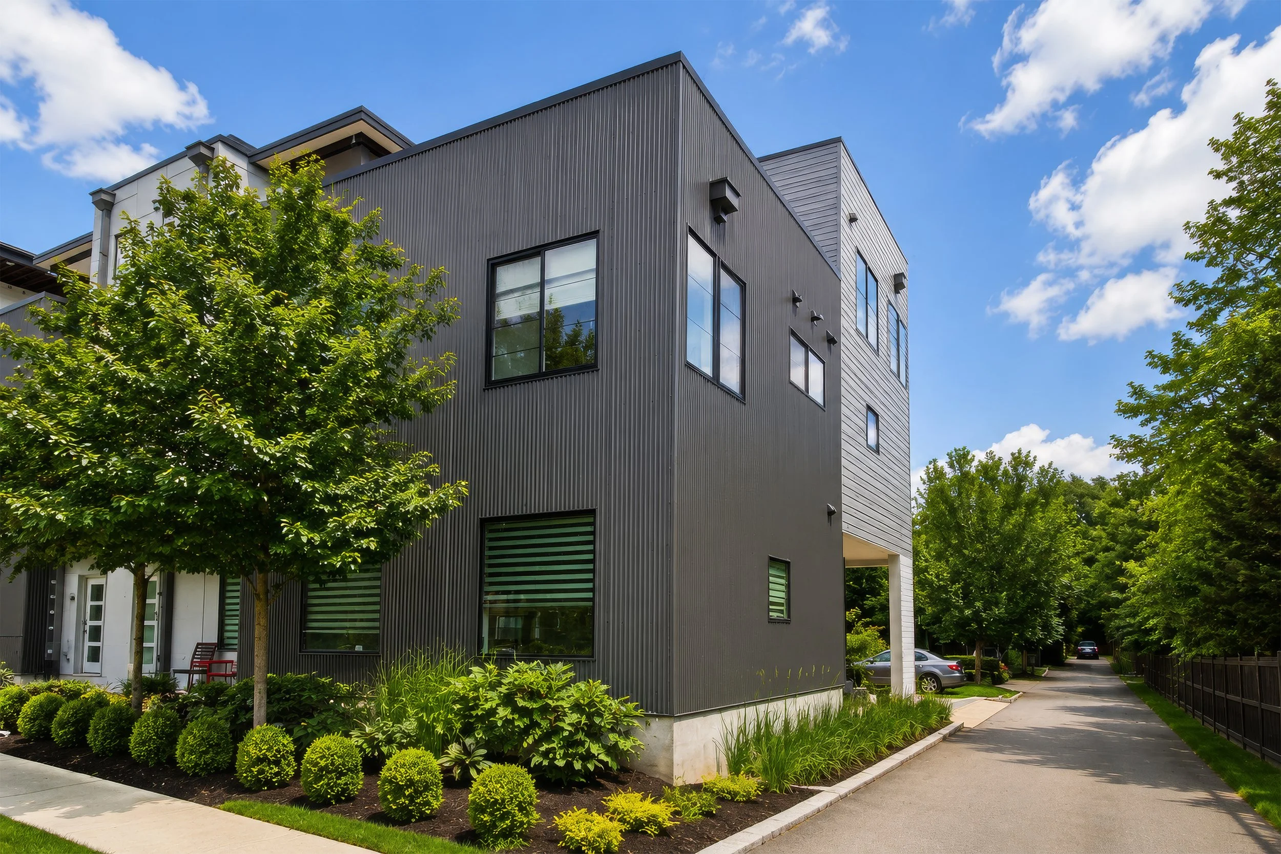 Modern multi-story apartment building with black exterior siding, large windows, and surrounding green trees and well-maintained landscaping under a blue sky with white clouds.