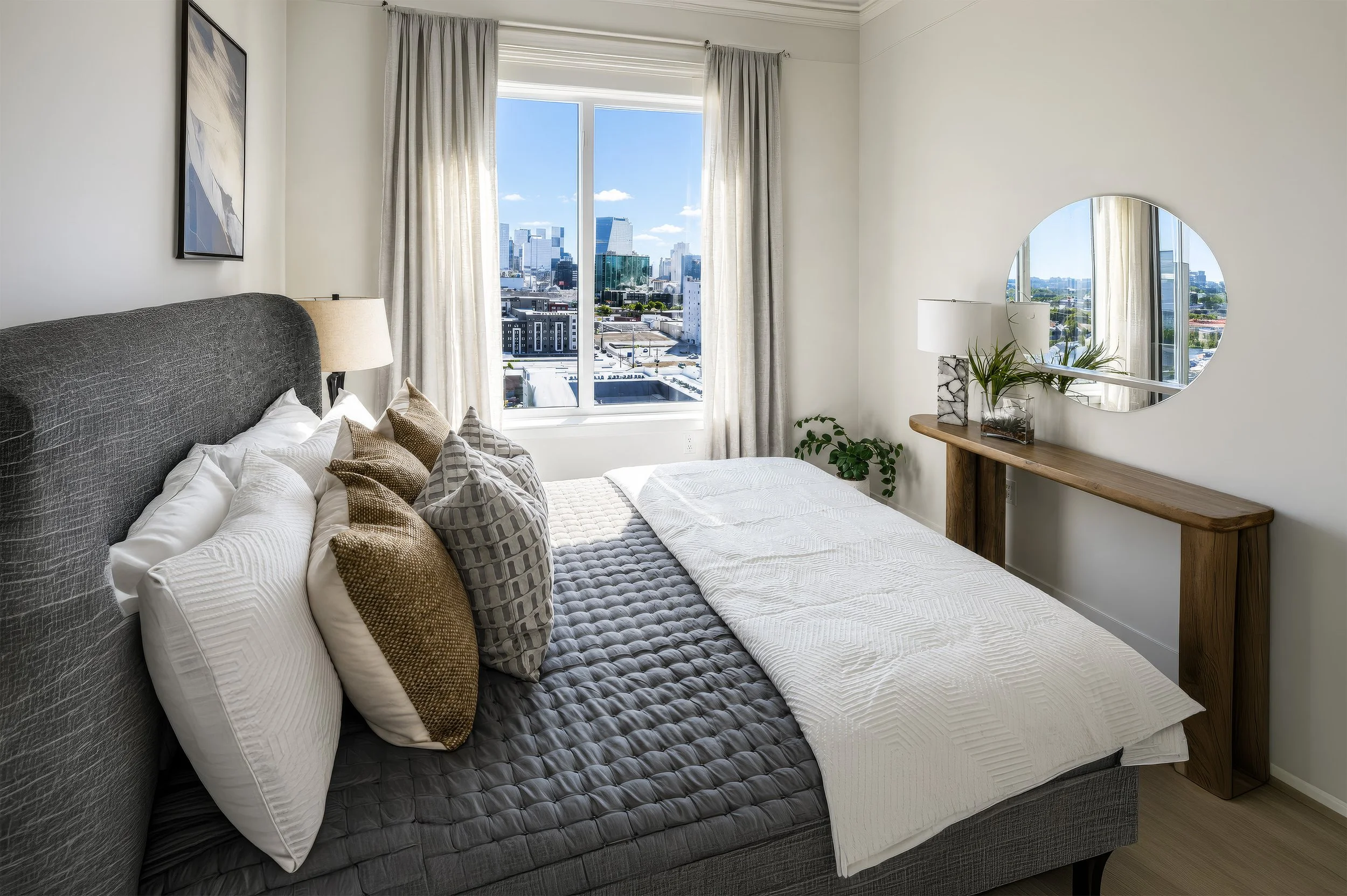 A bedroom with a large bed with white sheets and multiple decorative pillows. There are nightstands with lamps on each side of the bed. A window with light-colored curtains shows a cityscape with tall buildings outside. A wooden console table with a 