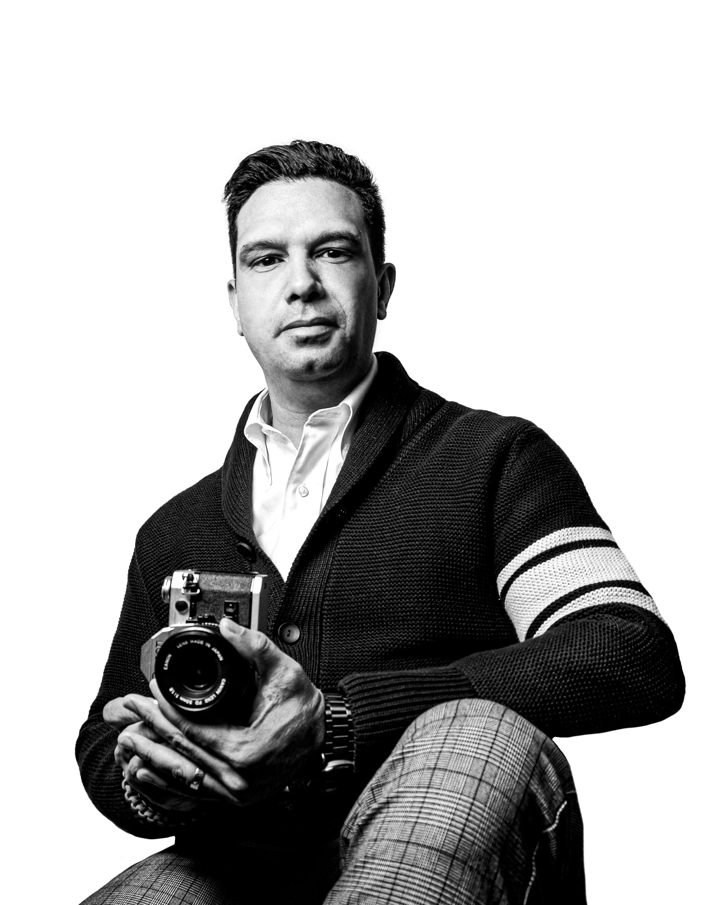 A man with dark hair taking a selfie in black and white, dressed in a sweater, collared shirt, and plaid pants, holding a vintage camera in his right hand, posing against a plain white background.