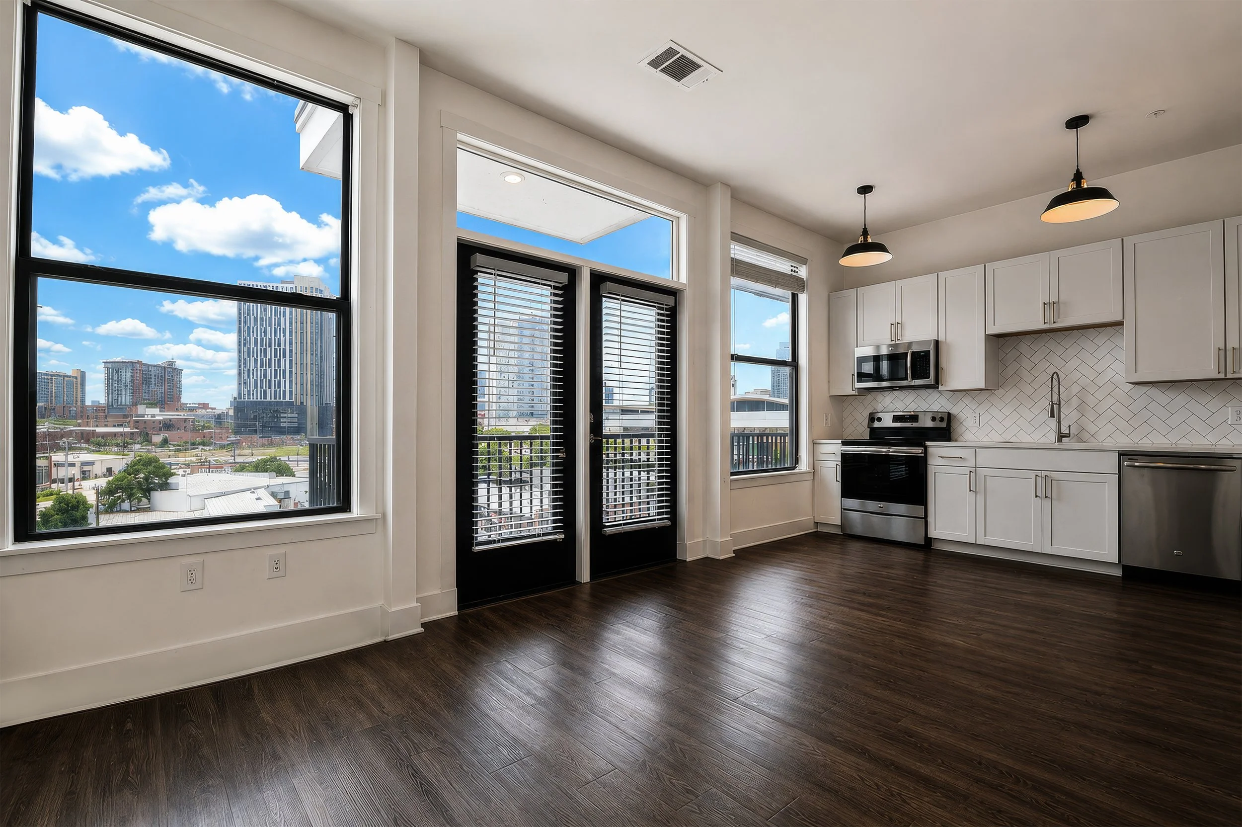 Bright modern kitchen with large windows showing cityscape, white cabinets, stainless steel appliances, dark hardwood floors, and pendant lights.