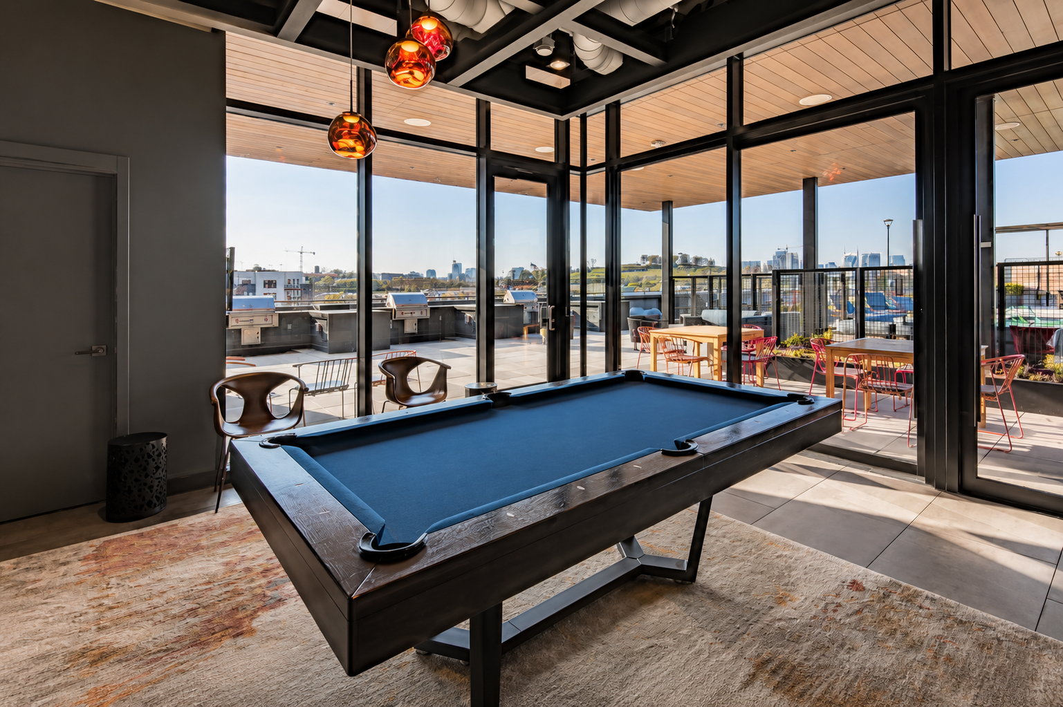 Indoor recreational room with a pool table, chairs, and large glass windows overlooking an outdoor patio with tables and chairs, cityscape view in the background.