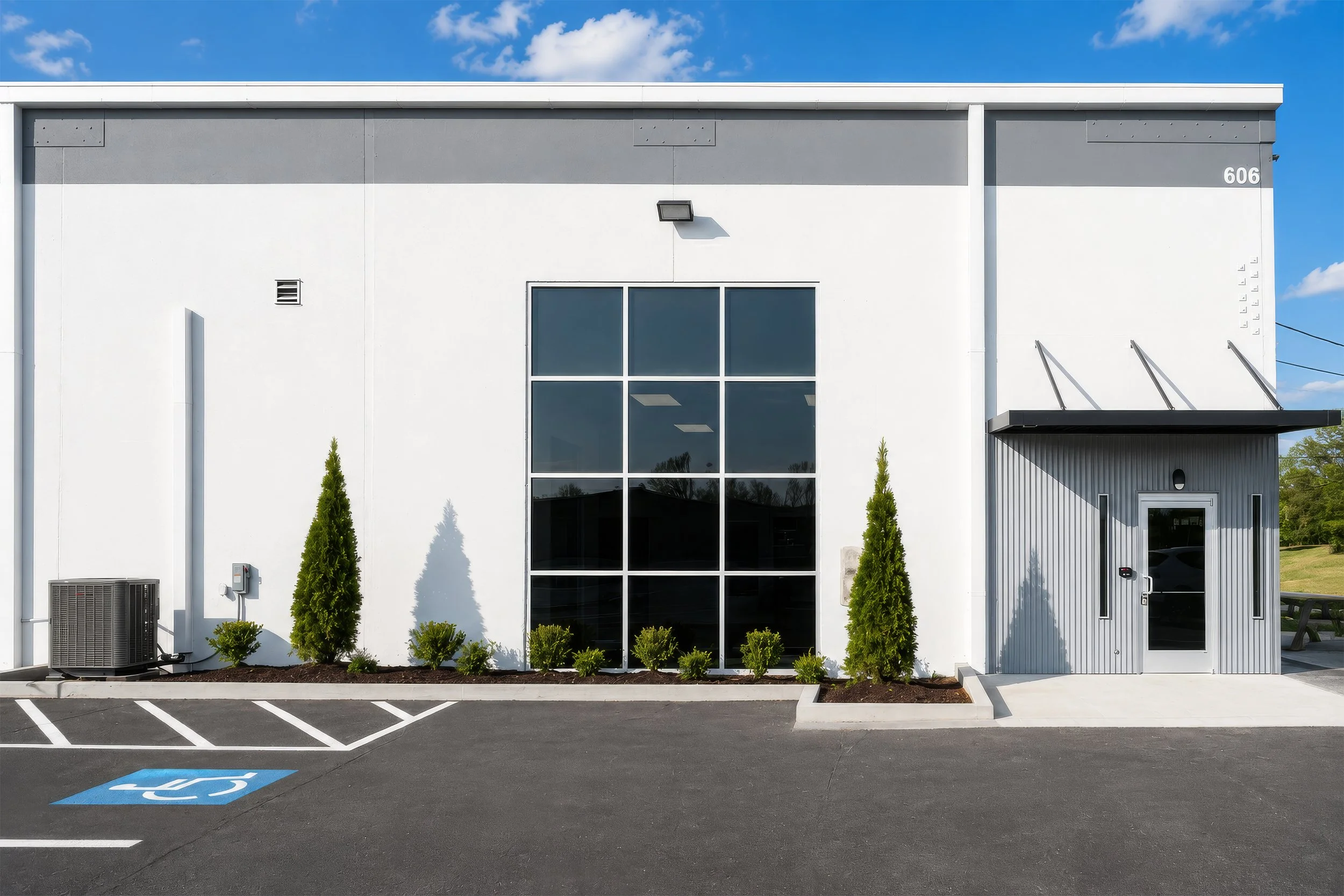 Front of a modern white commercial building with large glass windows, surrounded by a parking lot with a handicapped parking space, green shrubs, and a clear blue sky.