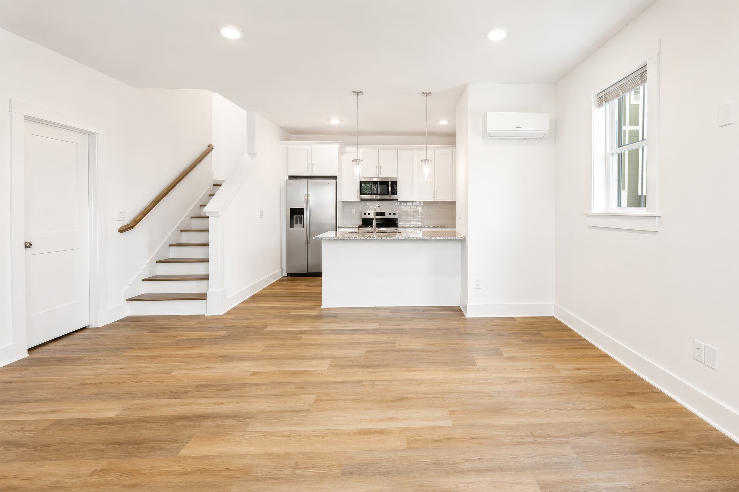A bright, white kitchen and living space with wooden floors, a staircase with wooden handrail, stainless steel refrigerator, microwave, oven, white cabinets, and a granite countertop. There is an air conditioning unit on the wall and a window with bl
