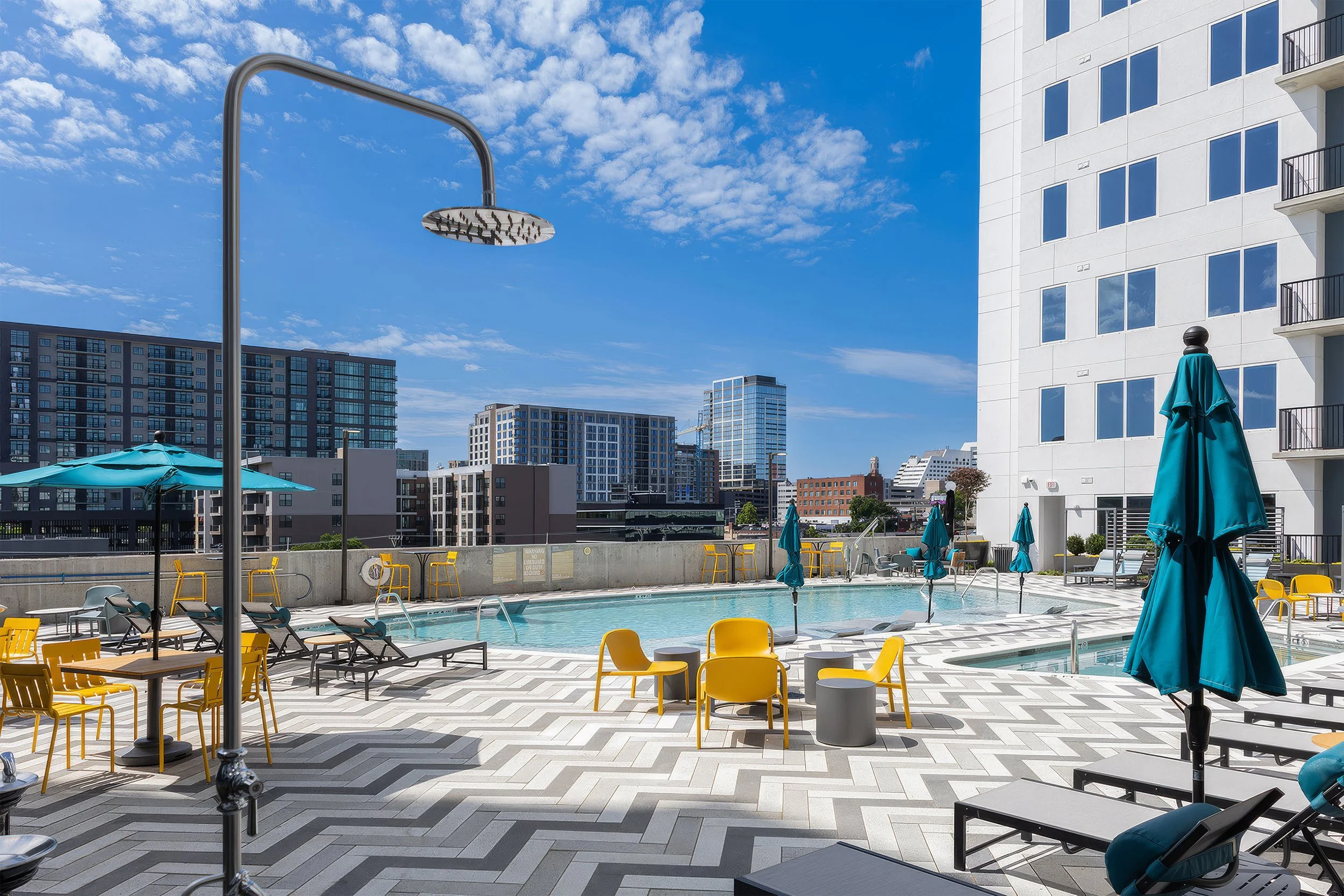 A rooftop pool area in a city with modern high-rise buildings, blue sky, and scattered clouds, featuring lounge chairs, blue umbrellas, and an outdoor shower.