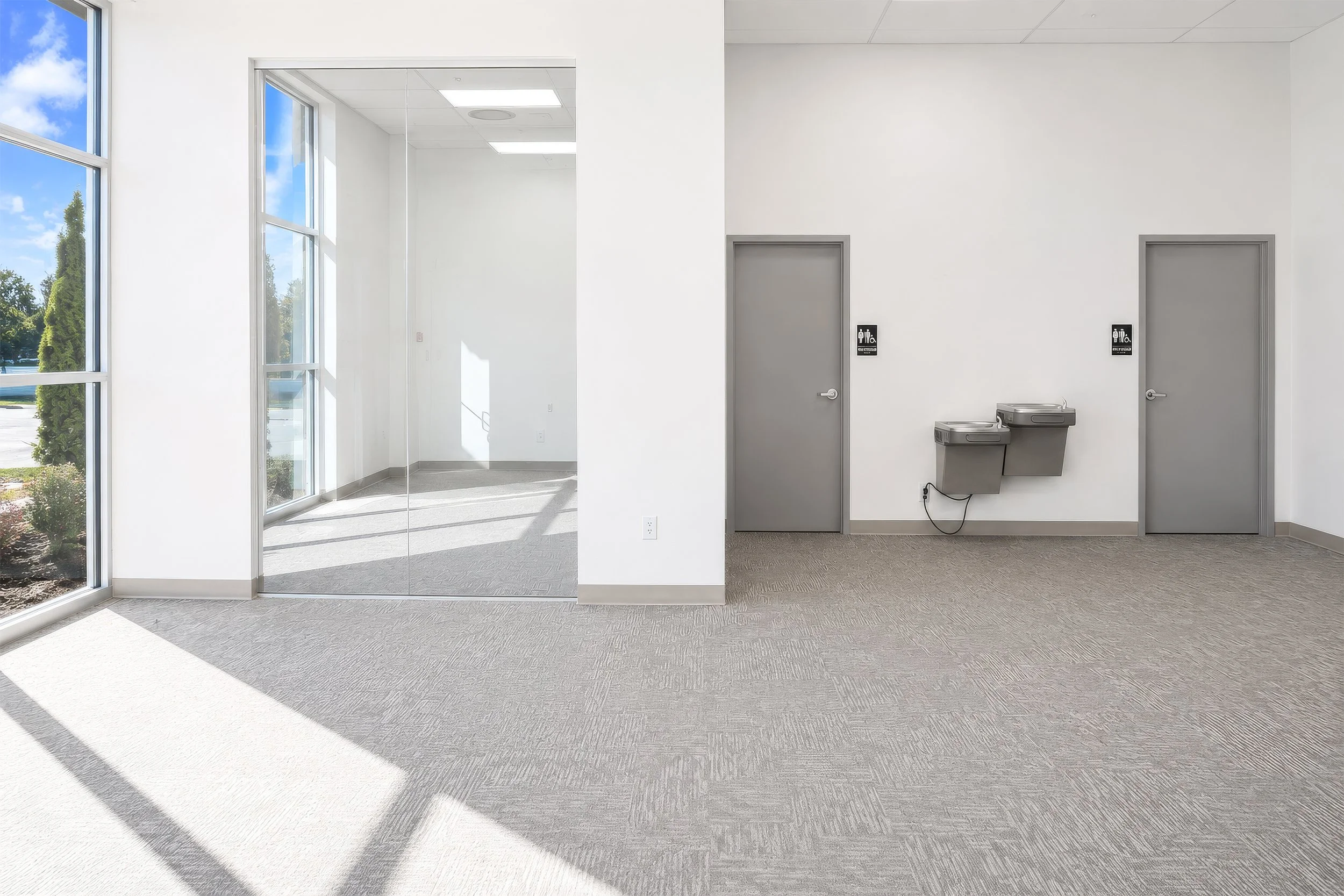 Interior of a modern building with large windows, gray doors labeled restrooms, and a drink fountain.