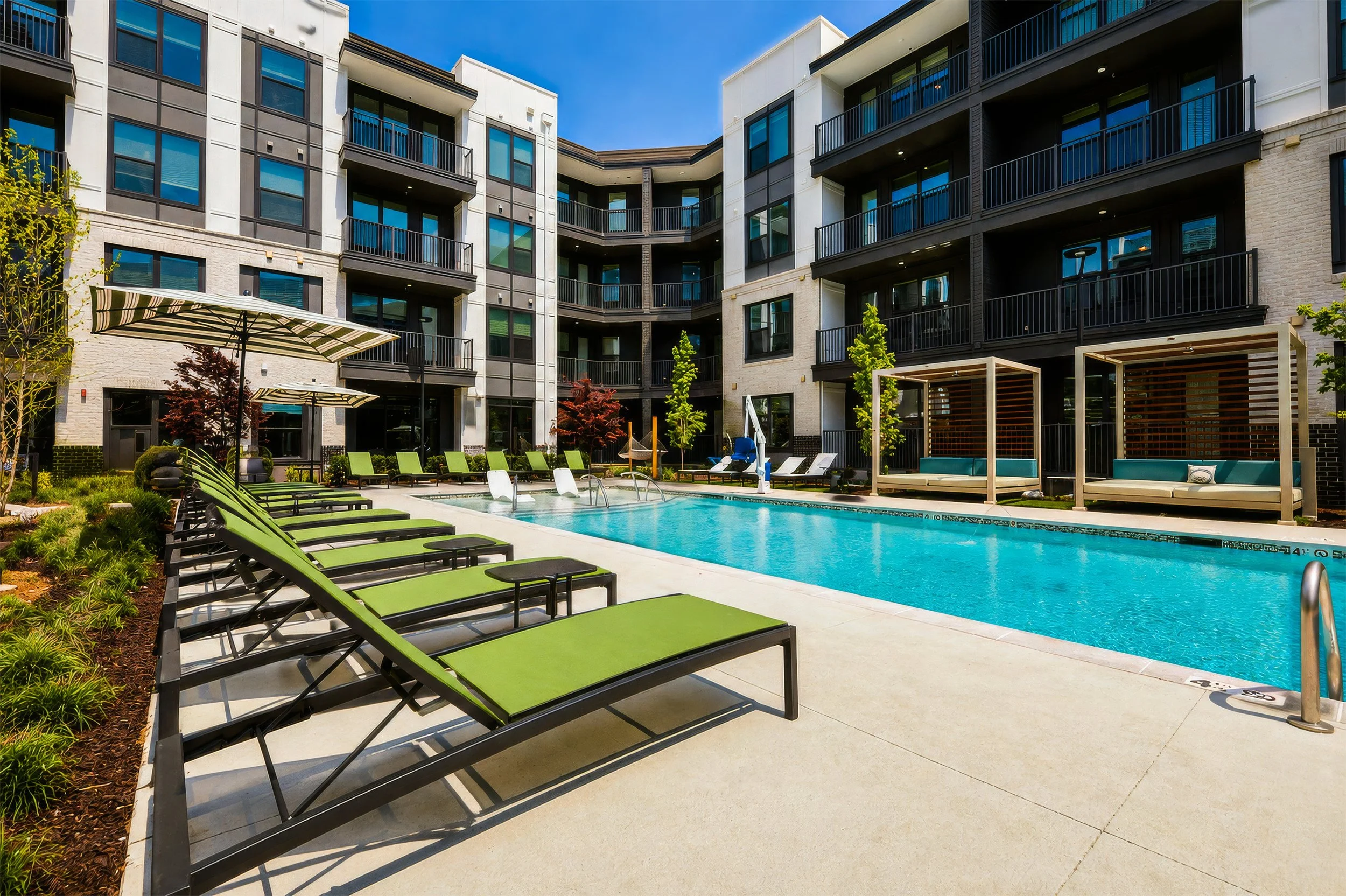 Outdoor swimming pool area surrounded by lounge chairs, umbrellas, and small trees in a modern apartment complex.