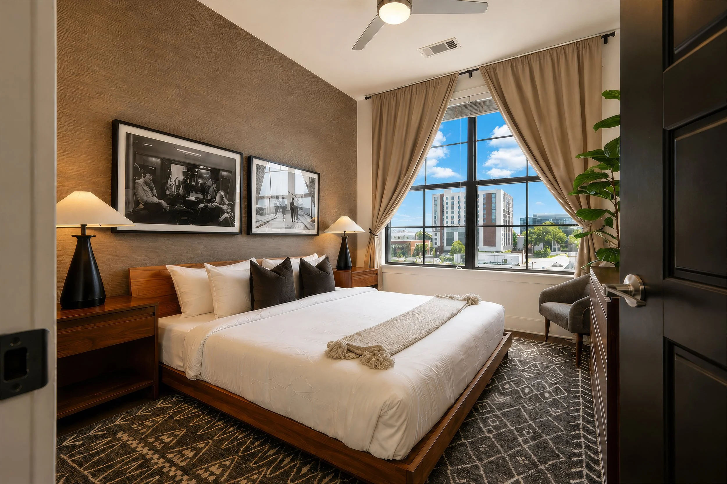 A bedroom with a large bed, dark wood nightstands, black and white framed photos on the wall, beige curtains, a large window showing a cityscape, a ceiling fan, and a potted plant.