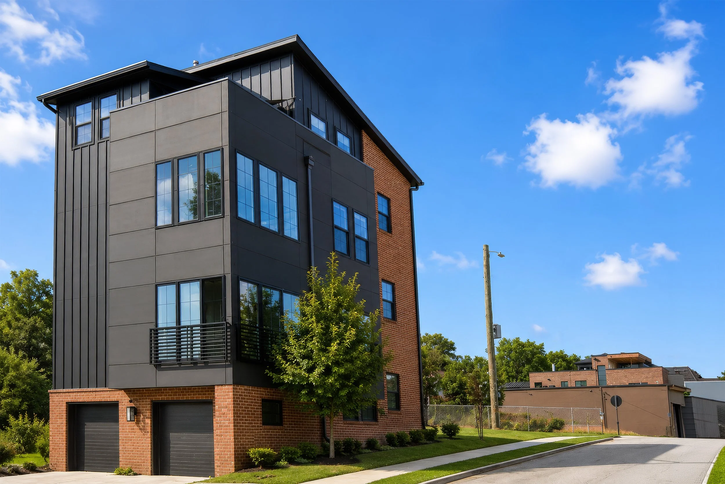 Modern multi-story residential building with a combination of brick, dark gray, and metal exterior, surrounded by green trees and a sidewalk, under a bright blue sky with few clouds.