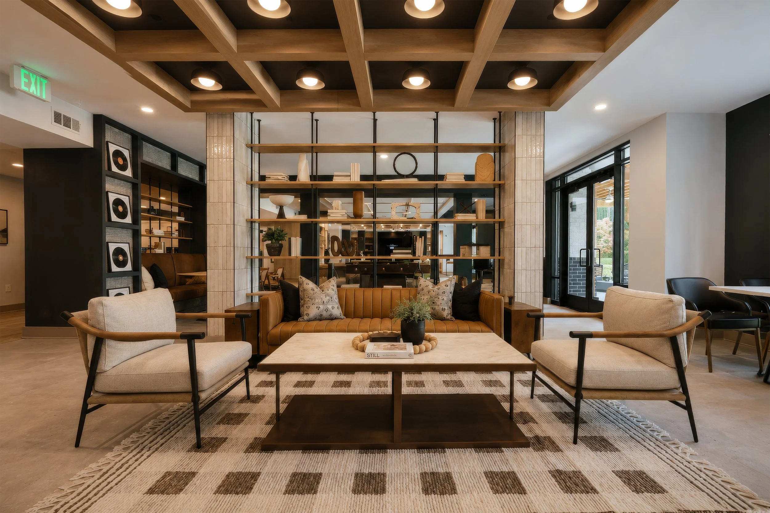 Contemporary living room with beige armchairs, a leather sofa, and a coffee table with a plant. A large open shelving unit with decorative objects and books separates the seating area from the background. The ceiling has wooden beams with recessed li
