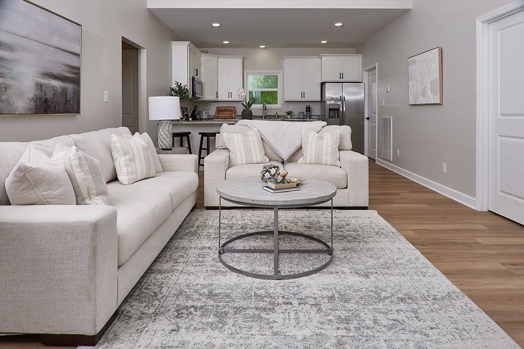 Living room with beige sofas, a round coffee table, a large area rug, and artwork on the walls, open to a kitchen area with white cabinets, stainless steel appliances, and a window.