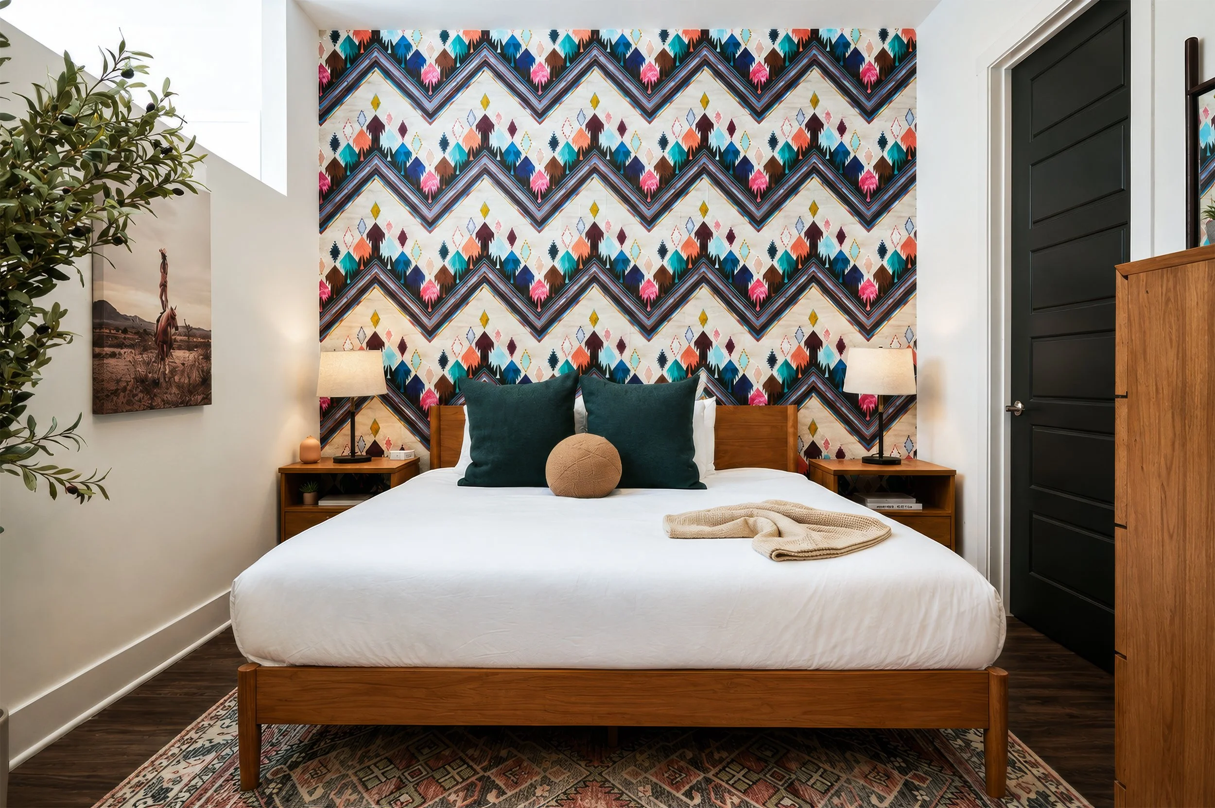 A bedroom with a patterned multicolored accent wall behind a wooden bed with white bedding, dark green pillows, and a tan round pillow. There are two wooden nightstands with lamps, a framed picture on the left wall, a plant on the left side, and a bl