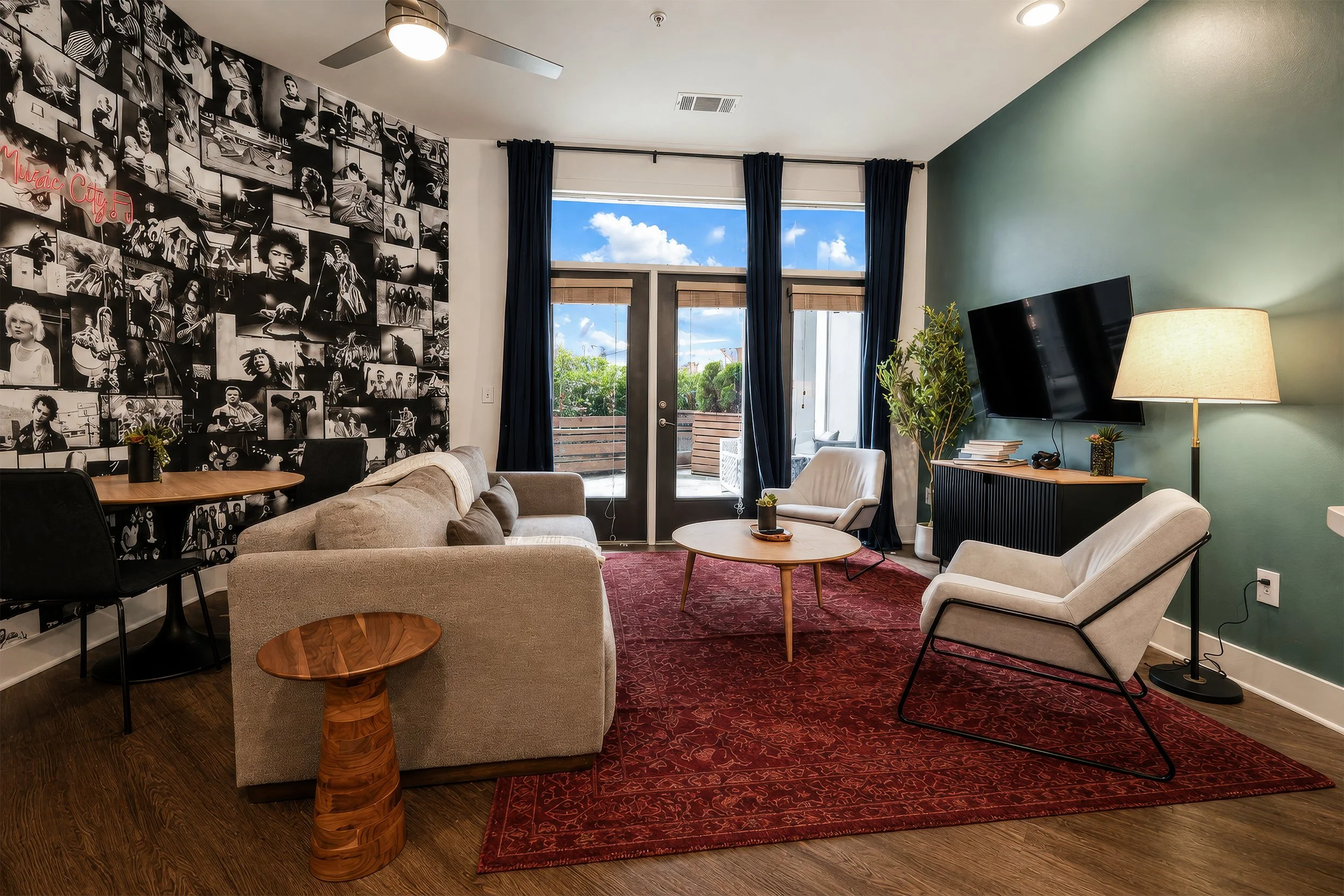 Living room with a black and white photo collage wall, blue curtains, a beige couch, a white armchair, a wooden coffee table with a small plant, a red patterned rug, a wall-mounted TV, a tall plant, a side table with books and a lamp, and a ceiling f