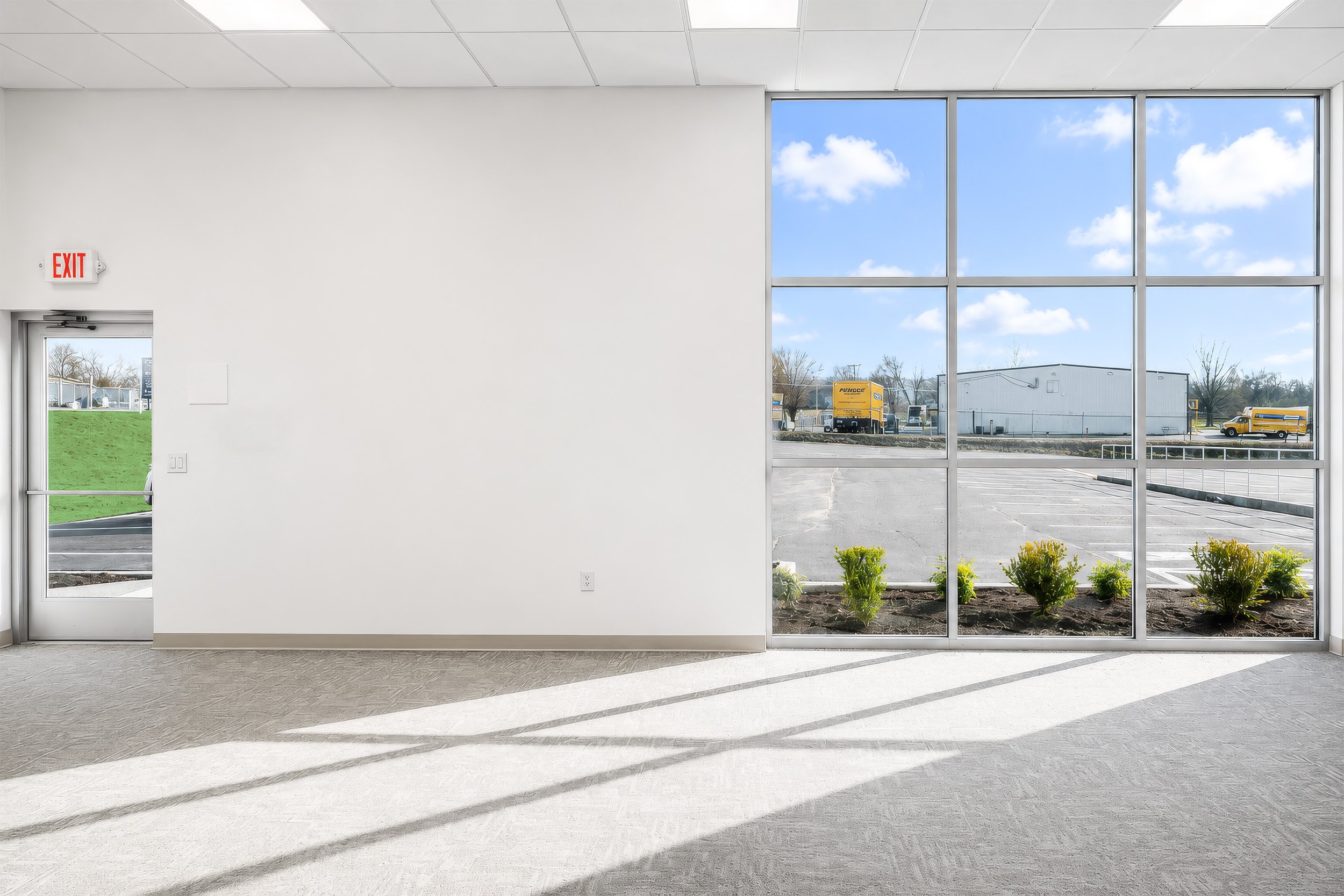 Inside view of a building with white walls and a large windowed wall showing a parking lot, yellow trucks, a white industrial building, and a partly cloudy sky.
