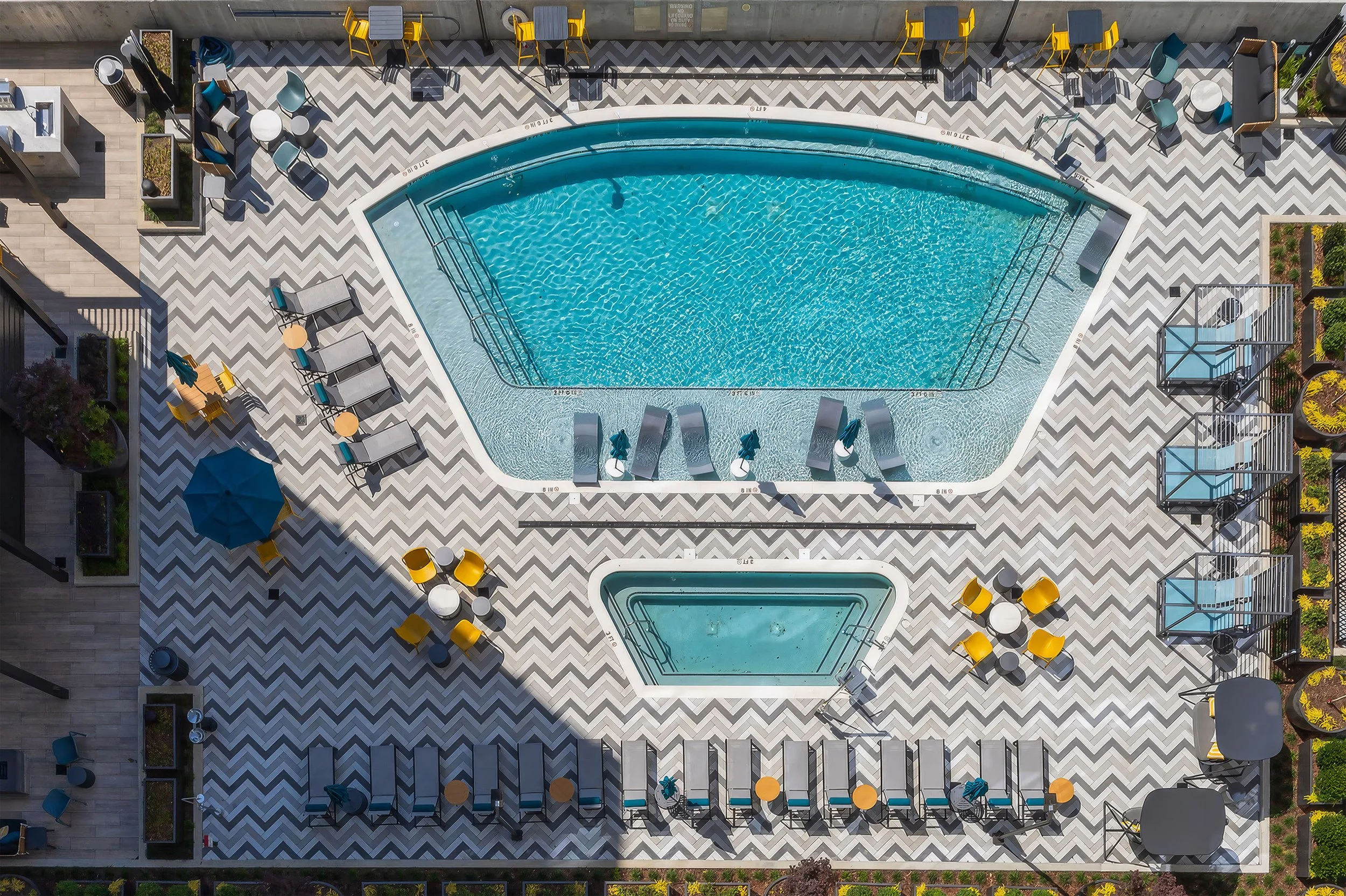 Aerial view of a pool area with lounge chairs, tables, chairs, umbrellas, and potted plants on a patterned pavement.