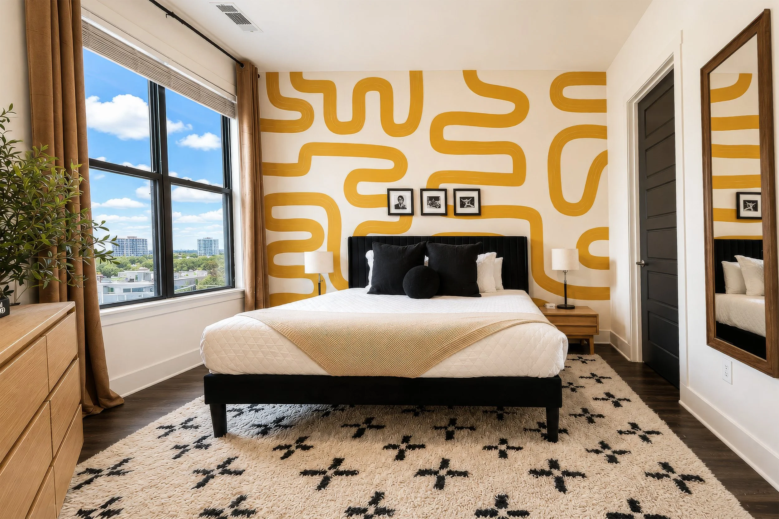 A modern bedroom with a large window showing a cityscape and blue sky with clouds. The room features a black bed with a textured white mattress, black and white pillows, two bedside tables with lamps, a wooden dresser, a plant, a black and white patt