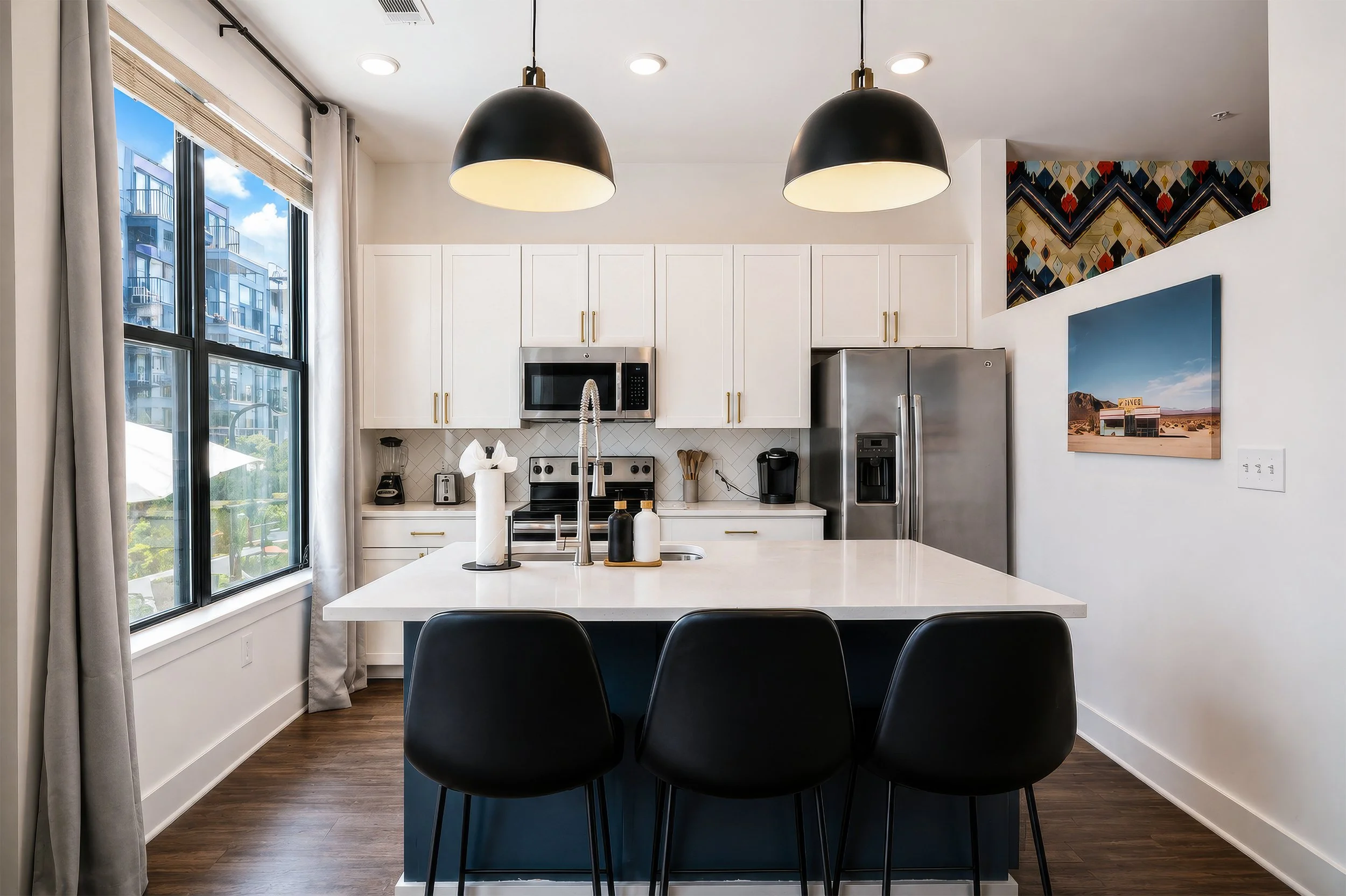Modern kitchen with large window, white cabinets, stainless steel appliances, black pendant lights, and a white countertop island with black chairs.