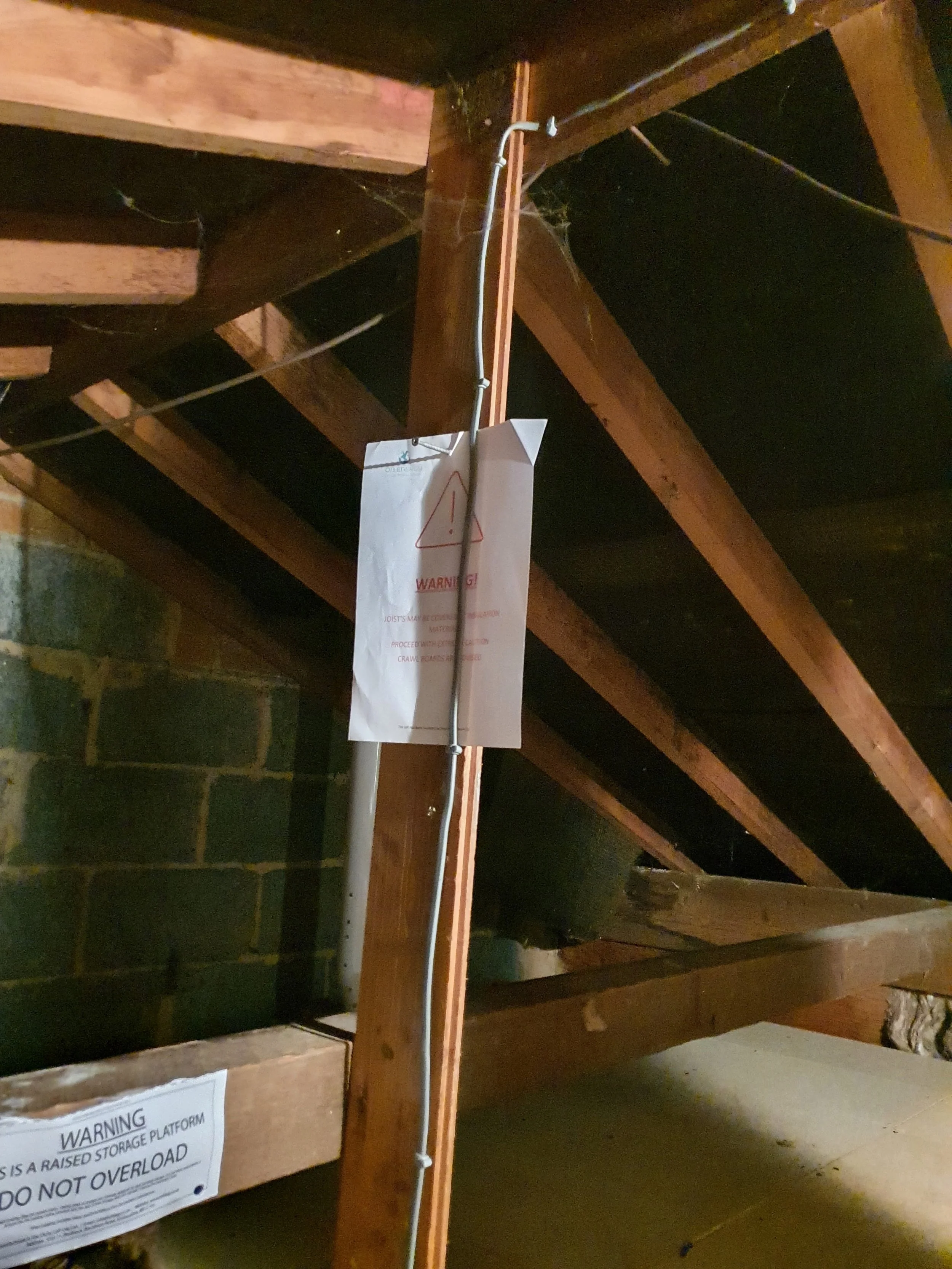 View of the underside of an attic or storage area with wooden beams, a metal conduit, and warning signs about storage platform and overload.