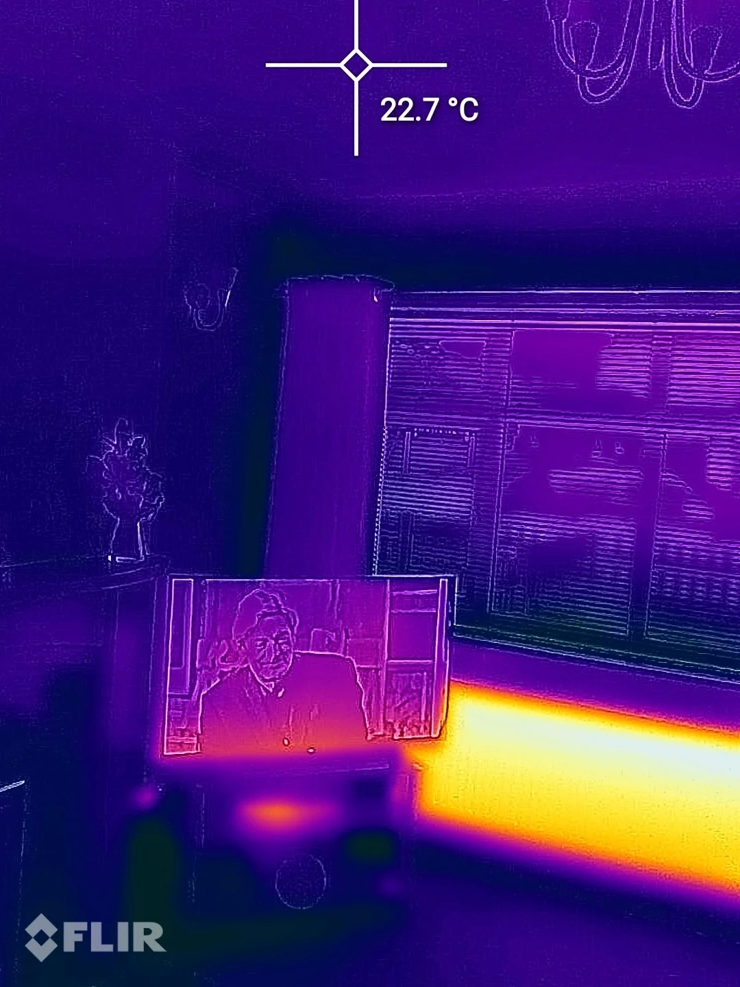 Thermal image of an indoor office setting showing a person on a screen, a temperature reading of 22.7°C, a clock, a plant, and furniture with varying heat signatures.