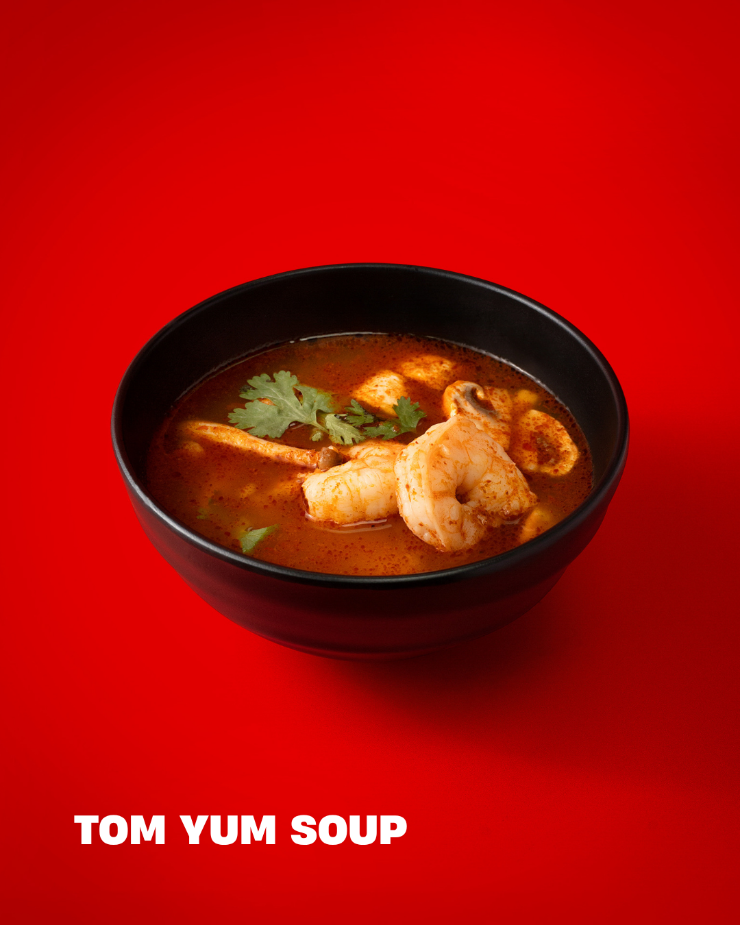 A black bowl of Tom Yum Soup on a red background with fresh shrimp, mushrooms, cilantro, and spices.