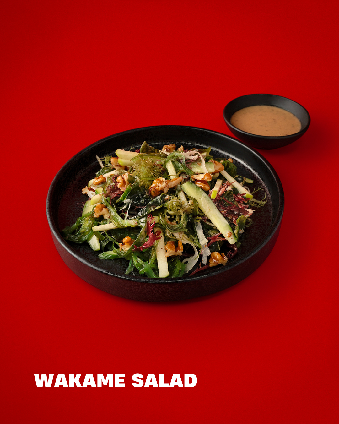 A black bowl filled with wakame salad, accompanied by a small black dish of dressing, on a red background, with the text 'WAKAME SALAD' at the bottom.