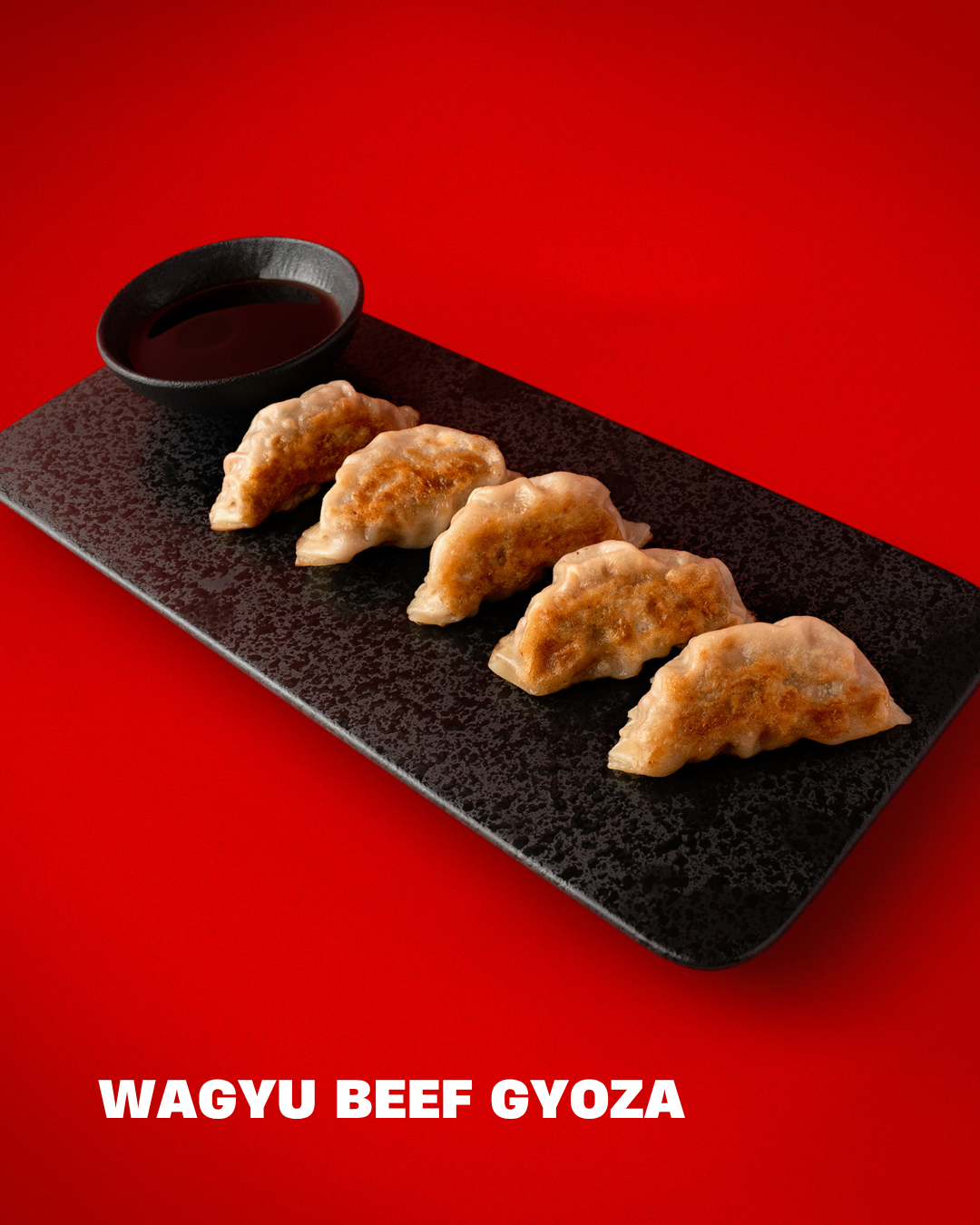 Six Wagyu beef gyoza dumplings on a black rectangular plate with a small bowl of dipping sauce, set against a red background.