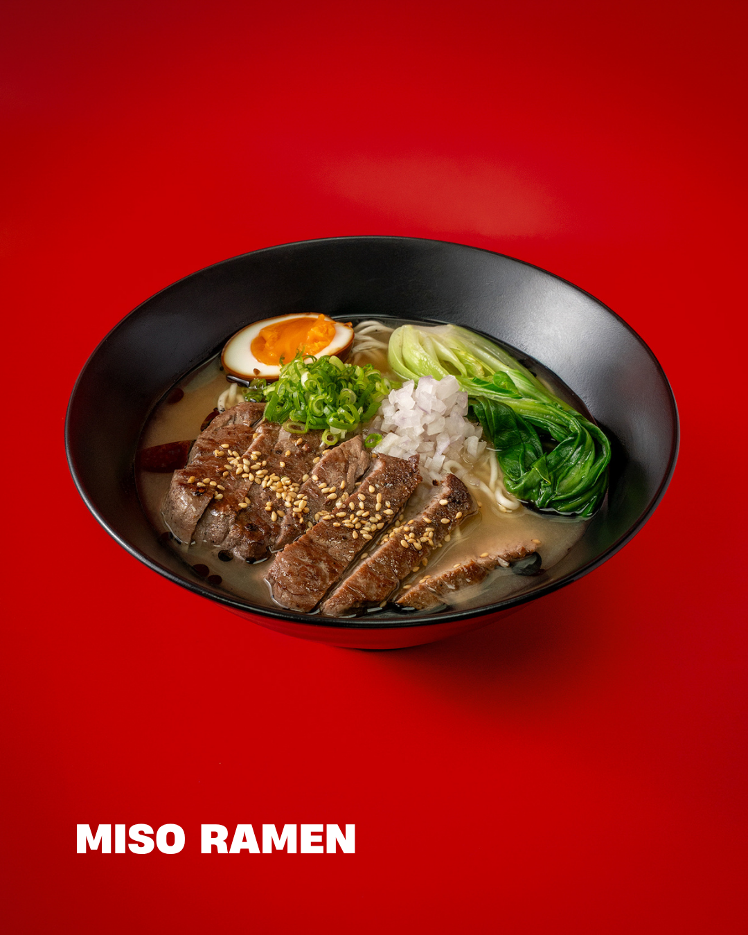 A bowl of miso ramen with sliced beef, soft boiled egg, green onions, chopped onions, bok choy, and noodles in broth, on a red background.