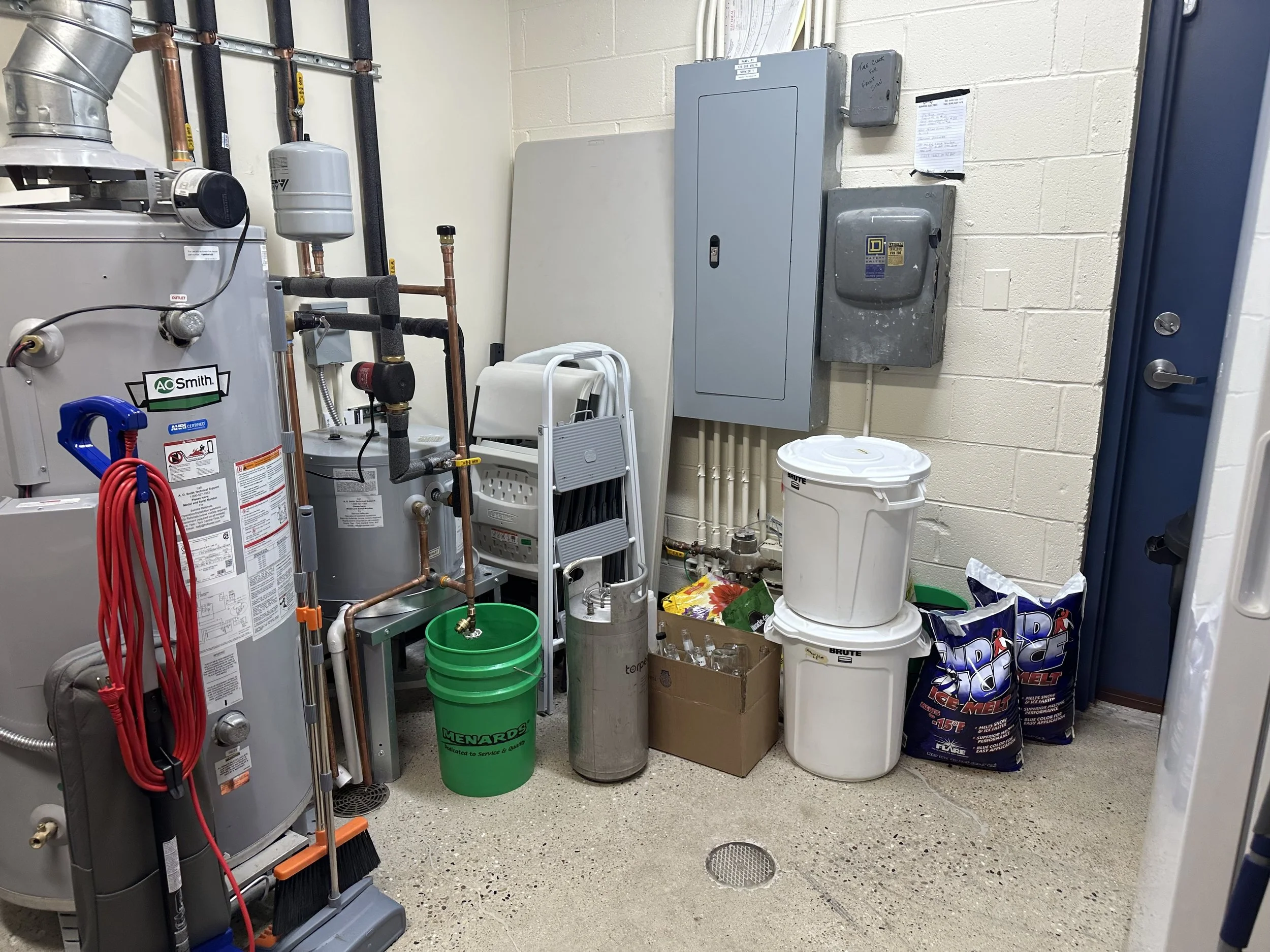 A utility closet containing a water heater, pipes, electrical panels, containers, and cleaning supplies.