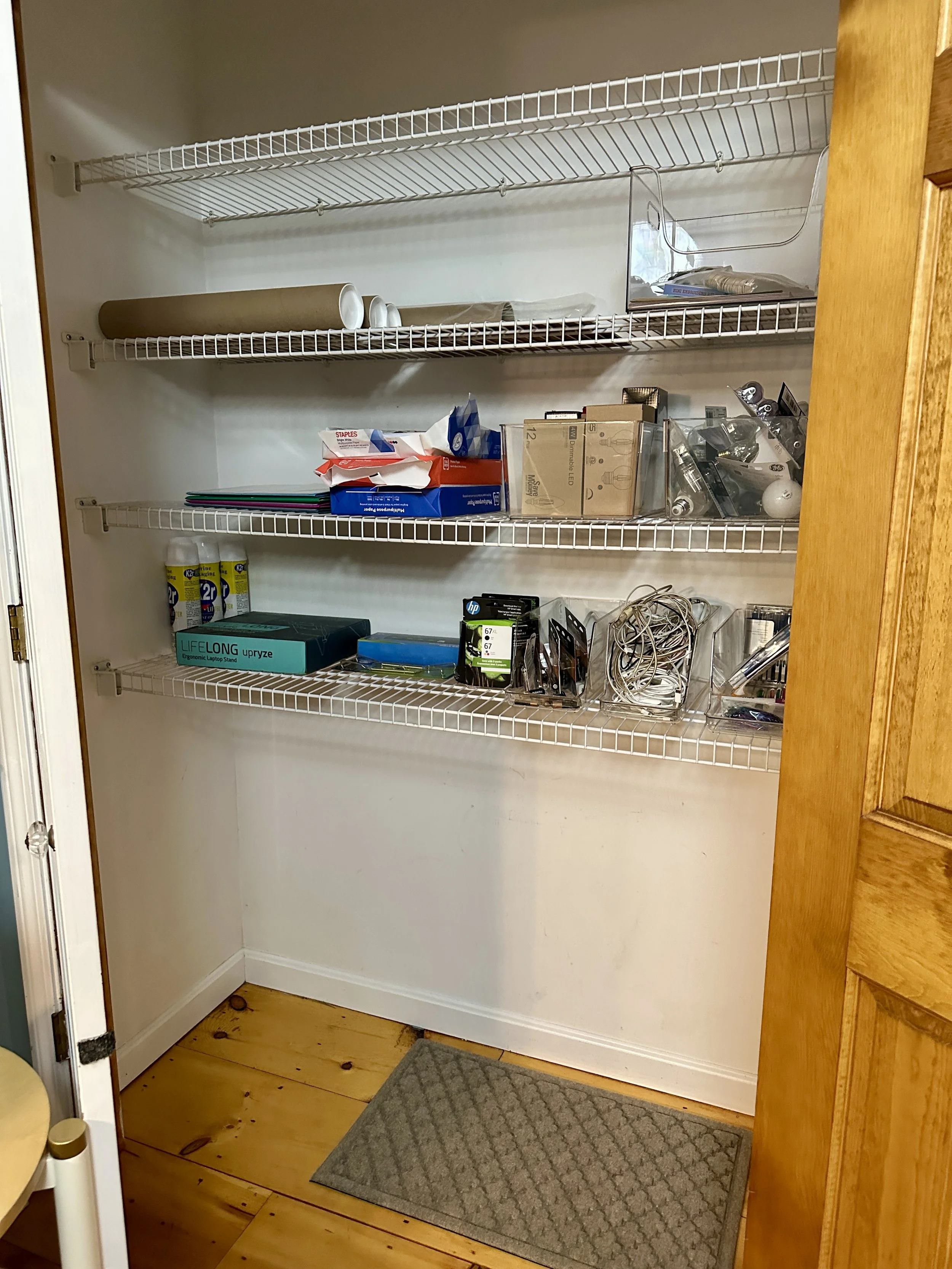 Empty pantry with white wire shelves, some office supplies, light bulbs, and miscellaneous stored items, with wooden flooring and a beige rug at the entrance.