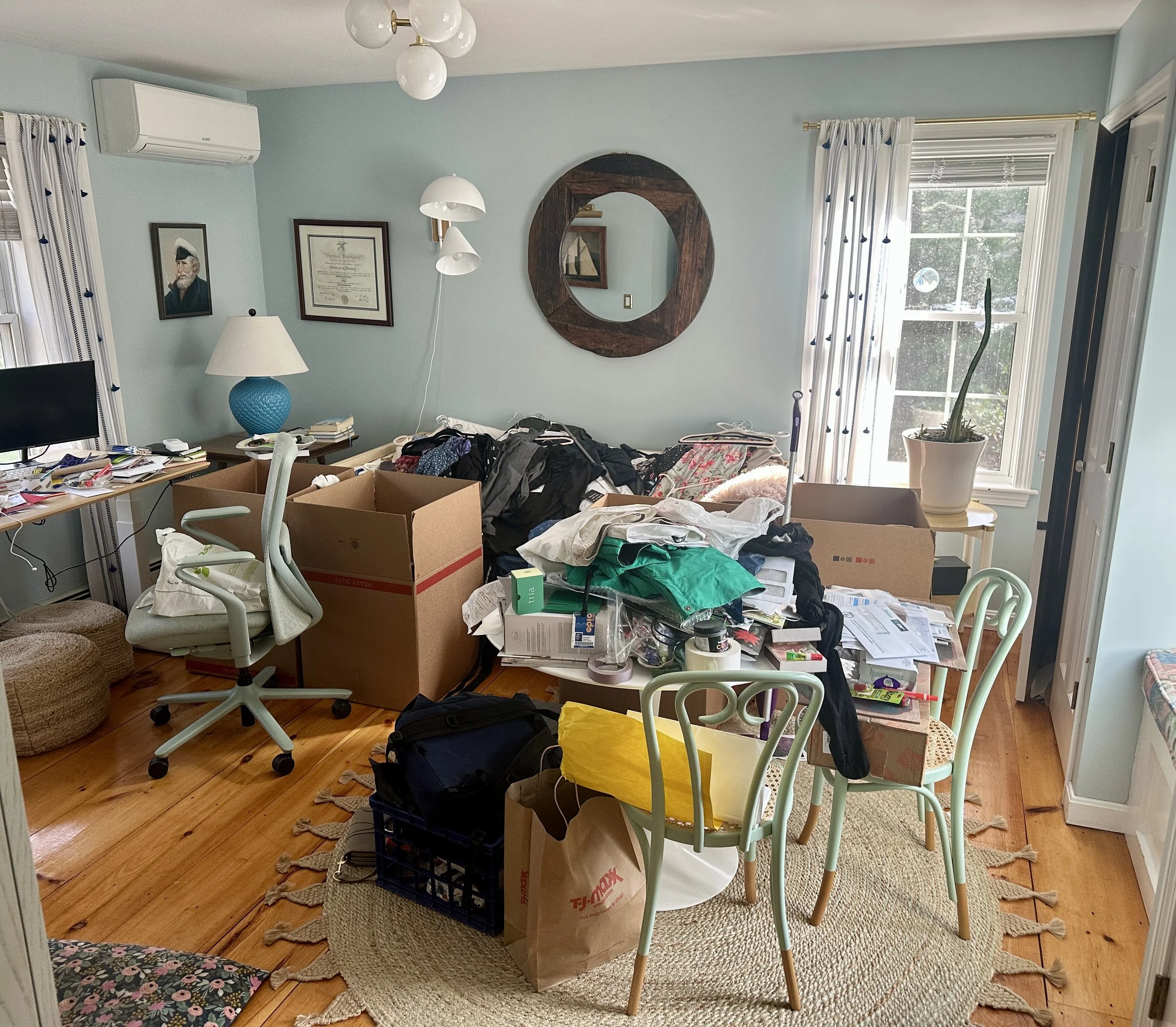 Living room with cluttered furniture and belongings, including boxes, chairs, a lamp, picture frames, and a plant, with sunlight coming through the windows.