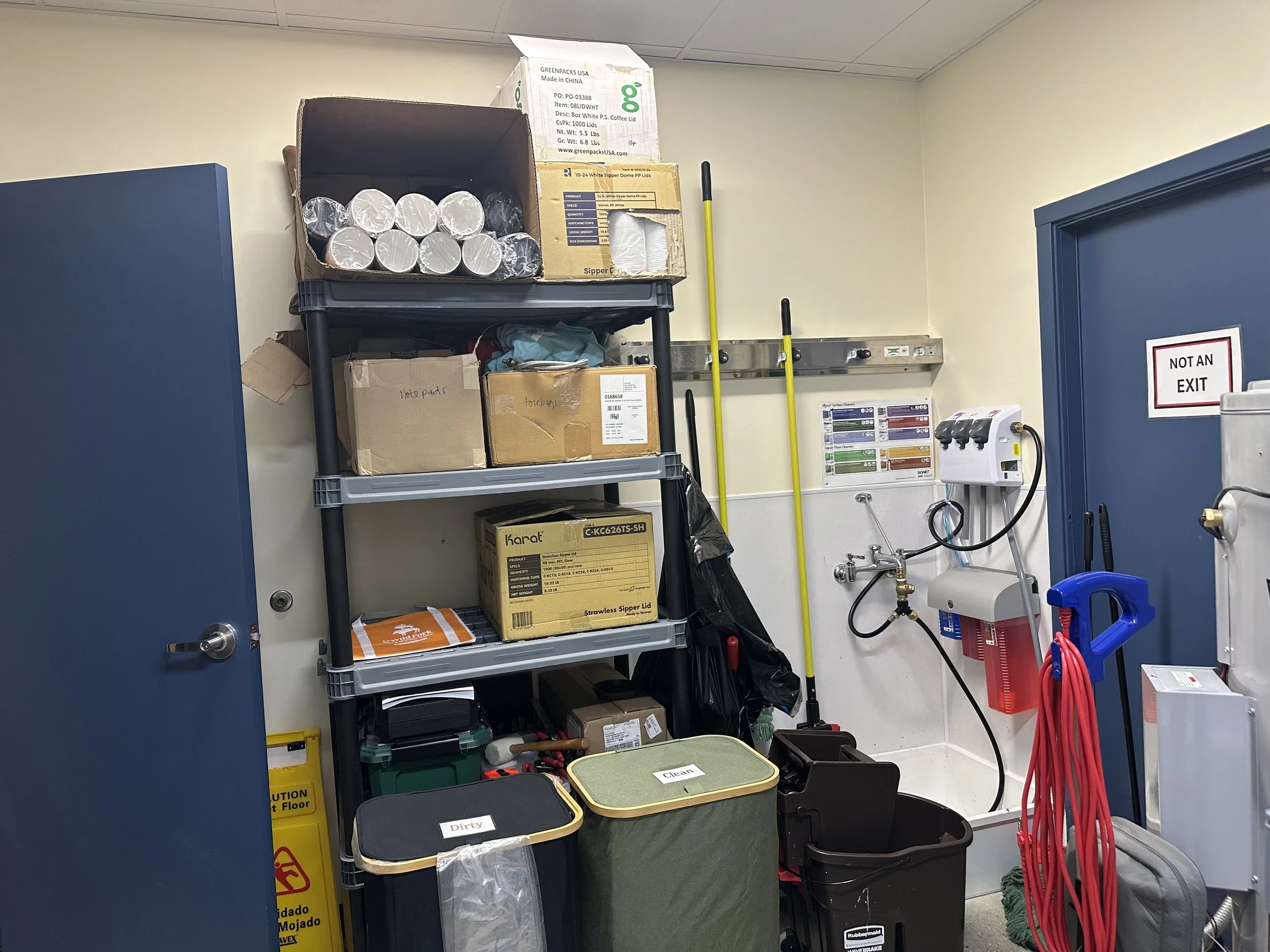 A storage area with shelves holding boxes and miscellaneous supplies, including cleaning supplies, cords, and equipment, next to a wall with a 'Not an Exit' sign.