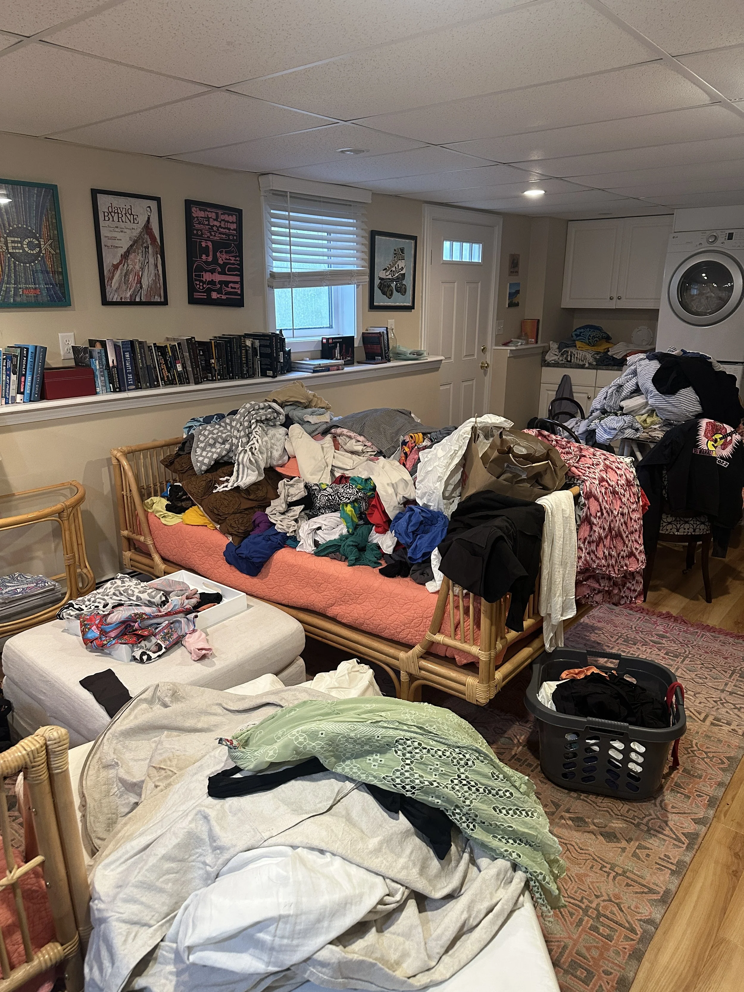 A cluttered living space with furniture, laundry, and household items scattered around. Clothes are piled on a bed and various surfaces, with laundry baskets and open drawers visible. The room has framed artwork on the walls, a window with blinds, an