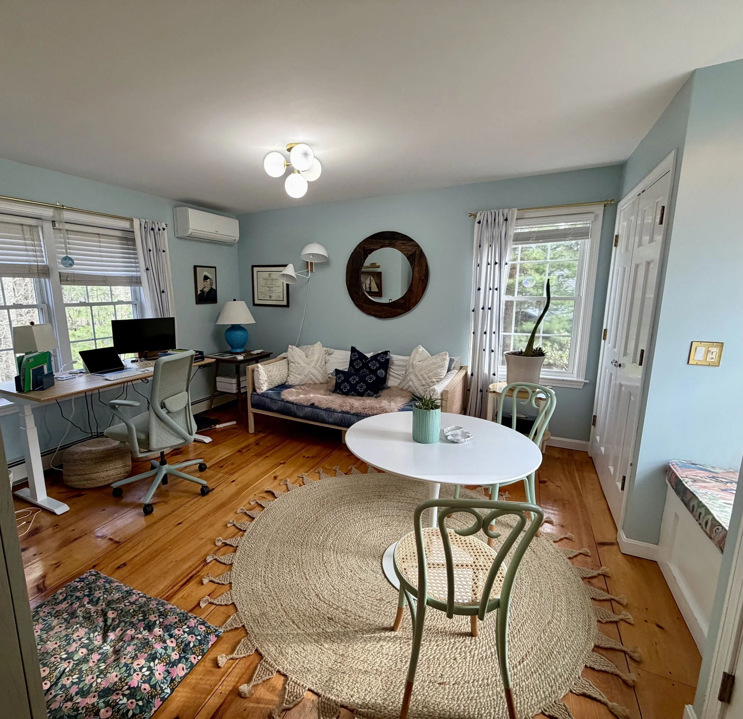 Living room with wooden floors, light blue walls, large windows with white curtains, a white round table with a blue pot and plant, beige and floral patterned rugs, a beige sofa with white and navy pillows, a wall mirror, framed pictures, a desk with a computer, and a white chair.